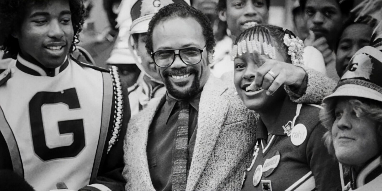 A lively group gathers, some in marching band uniforms, smiling and looking around. At the center stands a man in glasses and a suit, embodying Quincy's legacy, surrounded by others in festive attire.