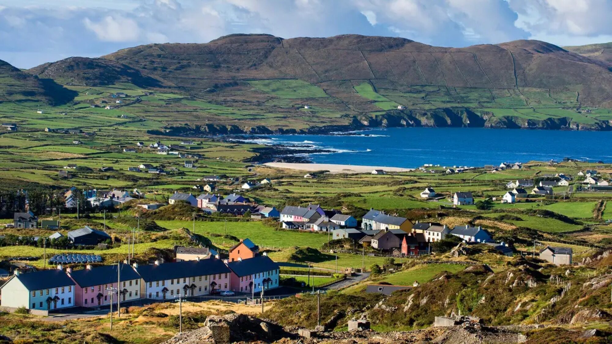 A coastal village with colorful houses, green fields, and hills overlooking the ocean under a partly cloudy sky.
