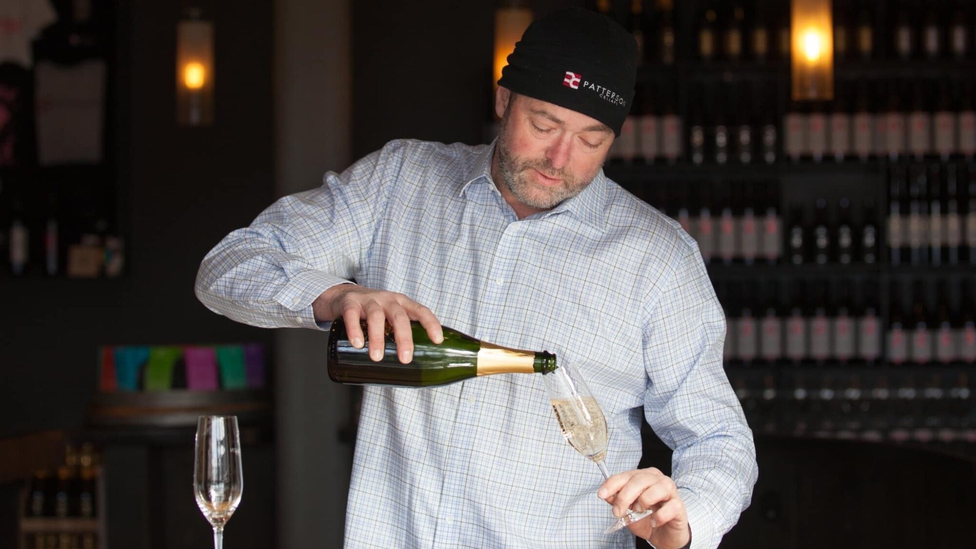 Man in a checkered shirt and beanie pouring champagne into a glass at a bar with wine bottles in the background.