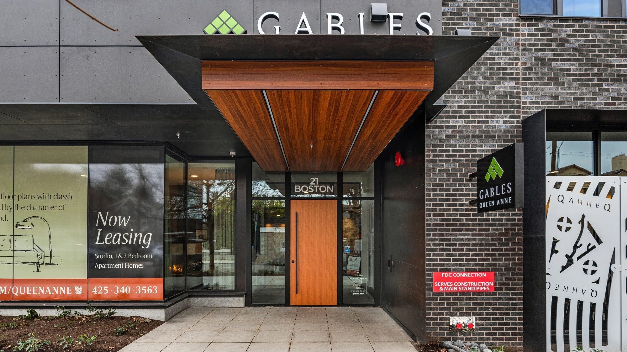 Entrance to a modern apartment building with a wooden door, signage for leasing, and construction notice. The building boasts a mix of brick and glass façade, subtly echoing Queen Anne architectural charm.