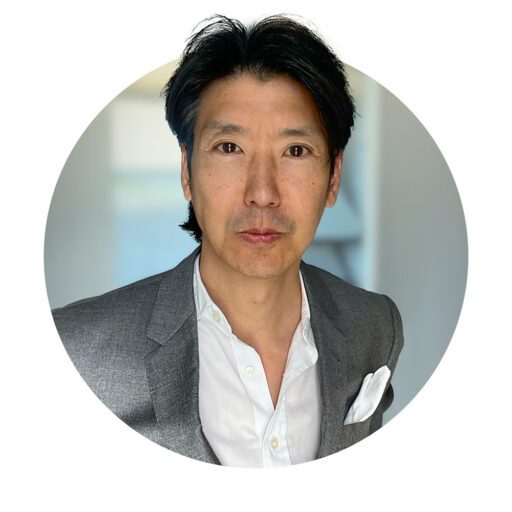 A man in a gray blazer and white shirt with a pocket square stands against a blurred indoor background.