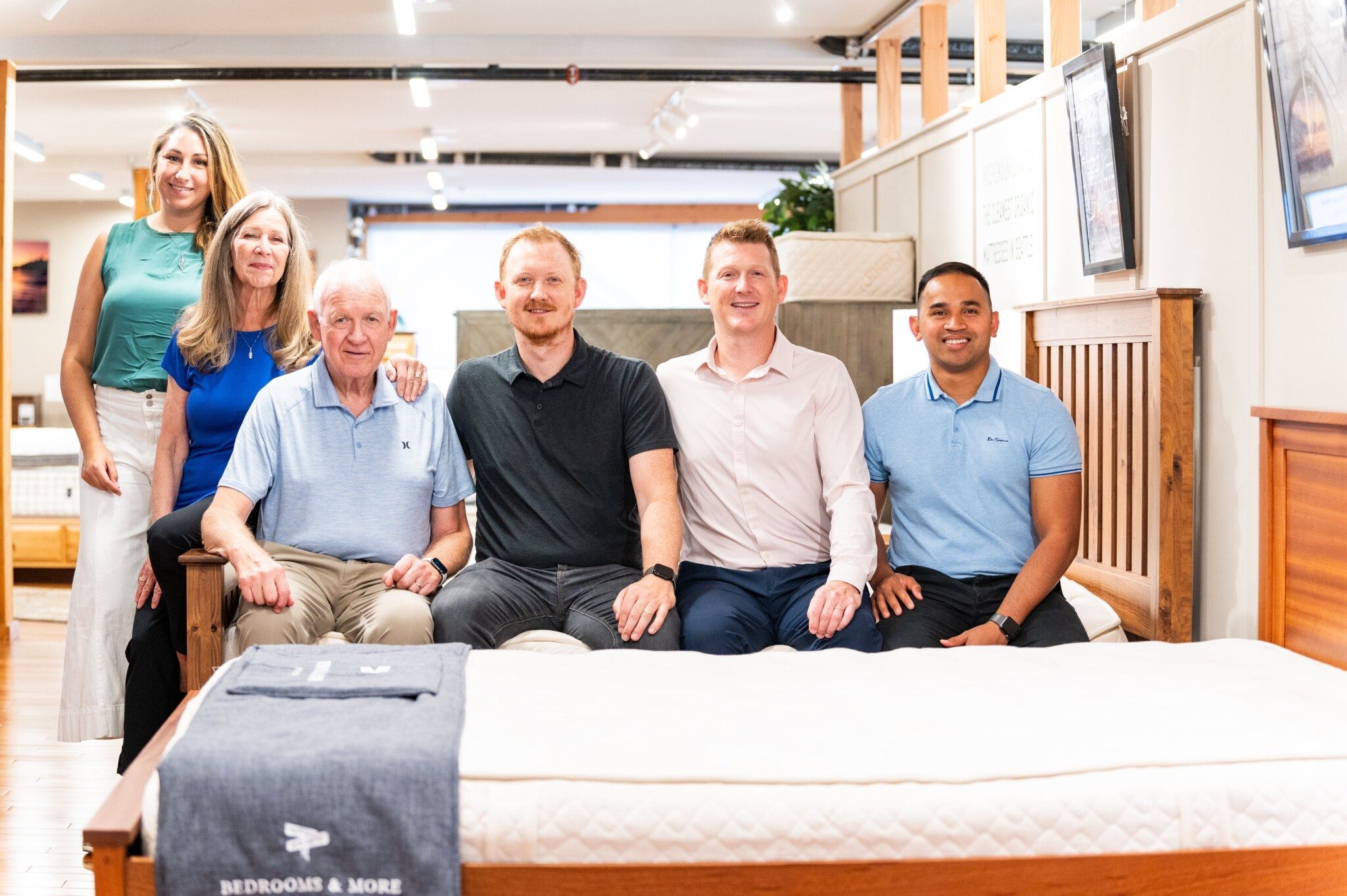 A group of six people sit and stand together in a furniture store, surrounded by wooden beds and decor items, imagining more perfect bedrooms in the background.