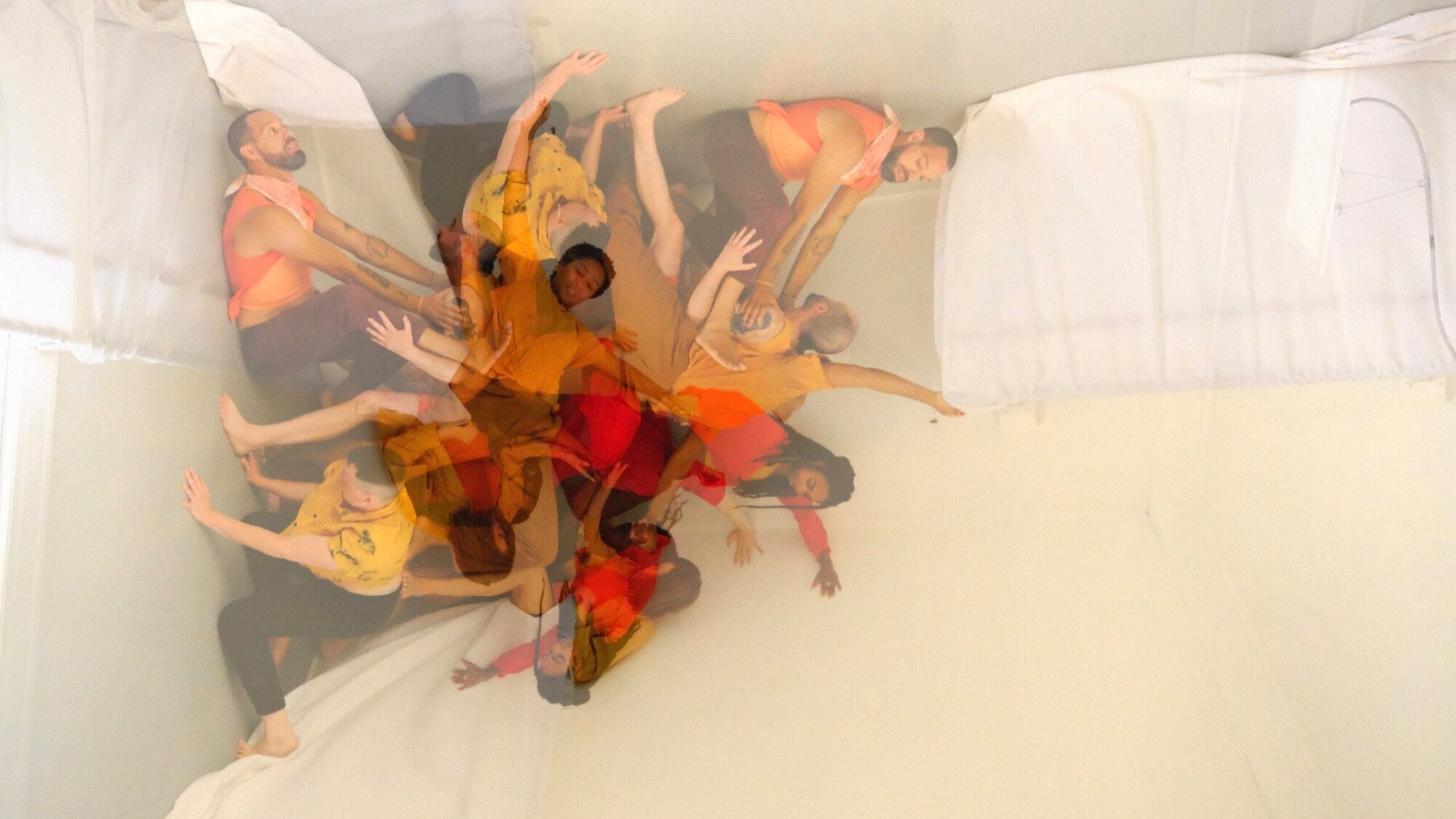 A group of people in colorful clothing are engaged in a dynamic, layered dance movement against a white background, creating a kaleidoscopic visual effect.