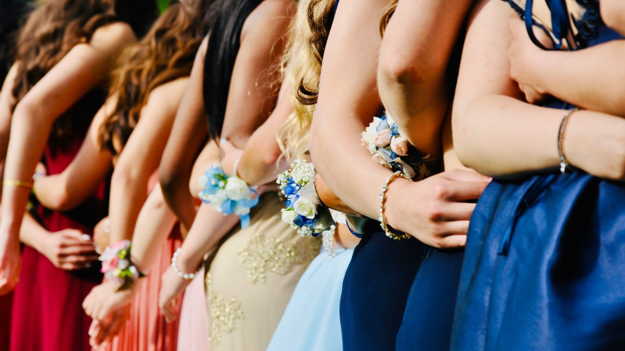 A line of people in formal dresses with corsages on their wrists, standing side by side and holding each other’s waists.
