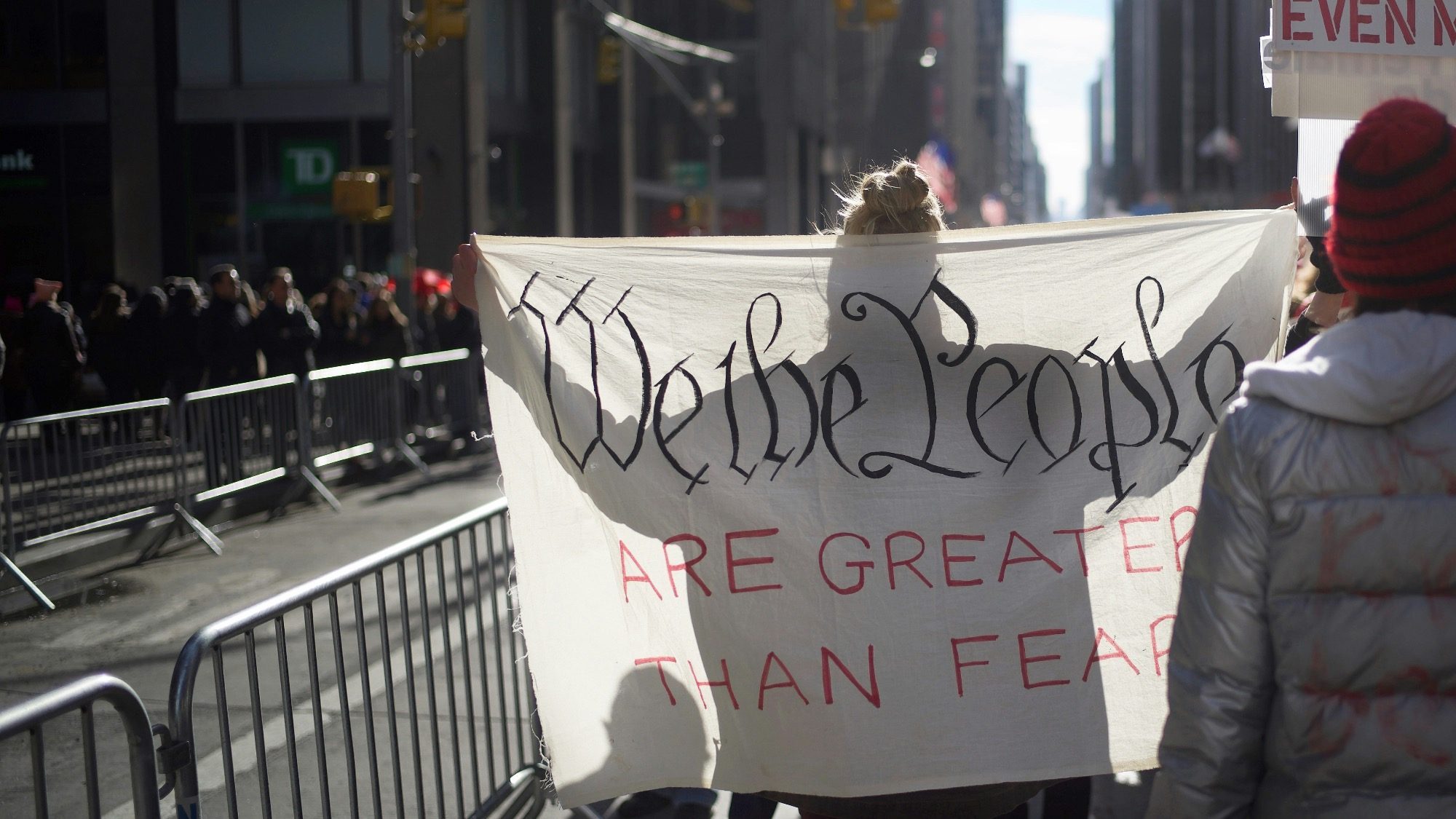 A person holds a sign reading "We the People Are Greater Than Fear" at a street demonstration, their voice resonating across all fifty states, with metal barricades and buildings in the background.