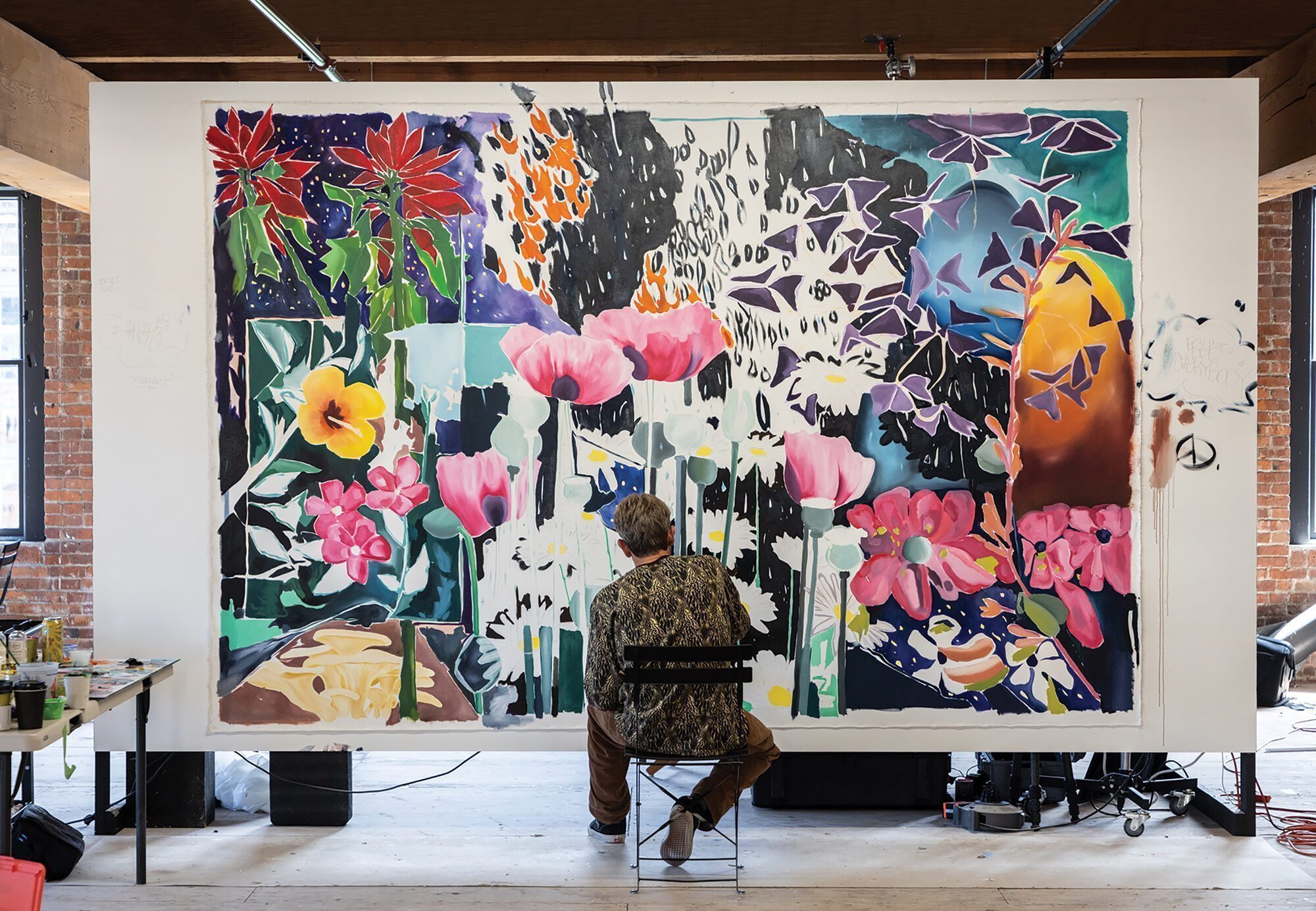 A person sits on a chair painting a large, colorful floral mural on a white wall in a spacious, well-lit studio near Pioneer Square Seattle's historic Hotel Westland, blending creativity and local charm for ARTXIV.