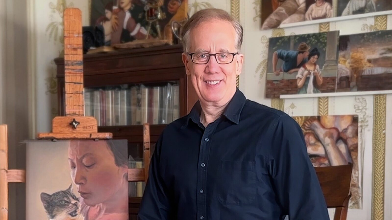 Former tech entrepreneur Andrew Conru stands smiling in an art studio, wearing glasses and a dark shirt. Behind him are various paintings and a bookshelf, with an easel displaying a portrait of a person with a cat. Conru recently announced the Seattle Prize Masters Fellowship.