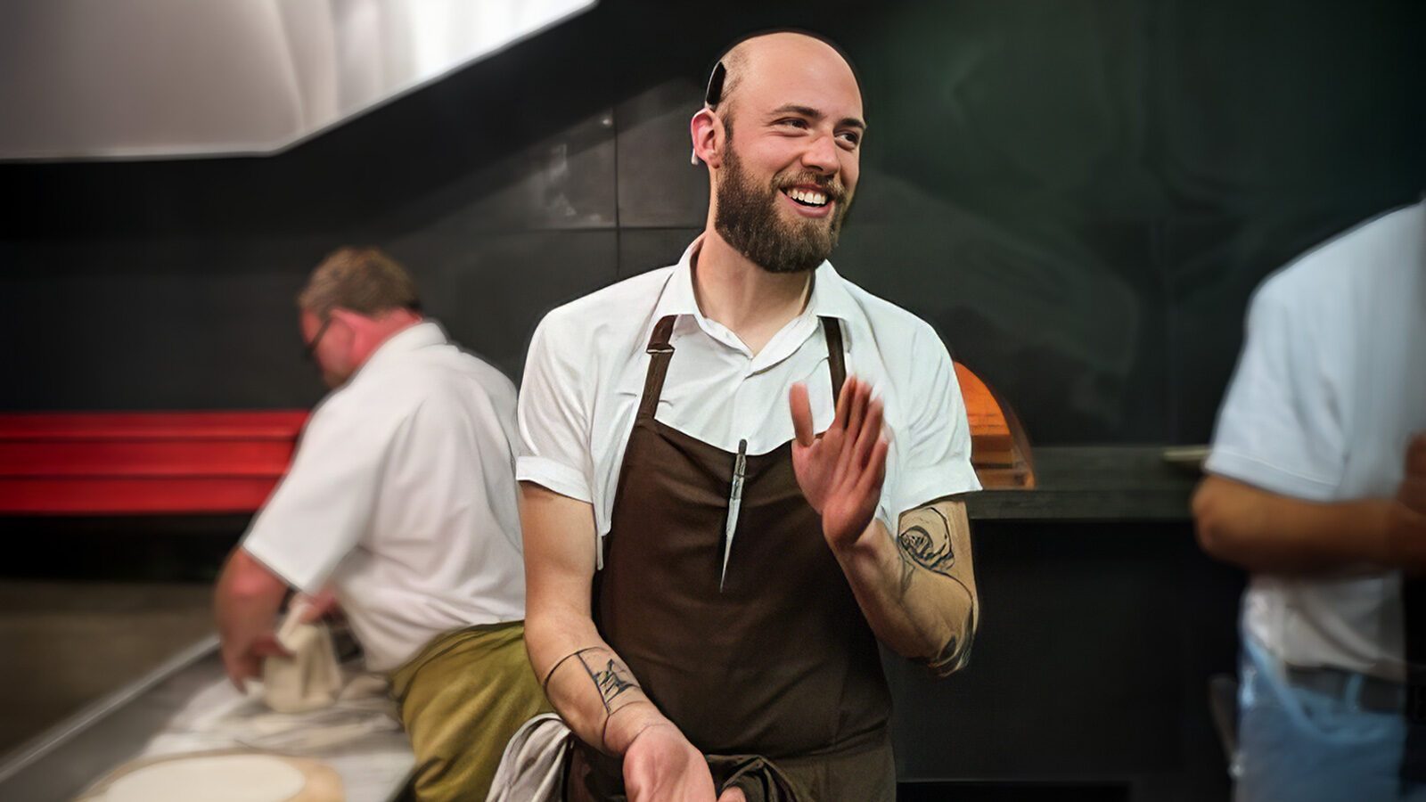 A bearded man wearing an apron and holding a cloth smiles while standing in a kitchen. Another person in the background is working on a task.