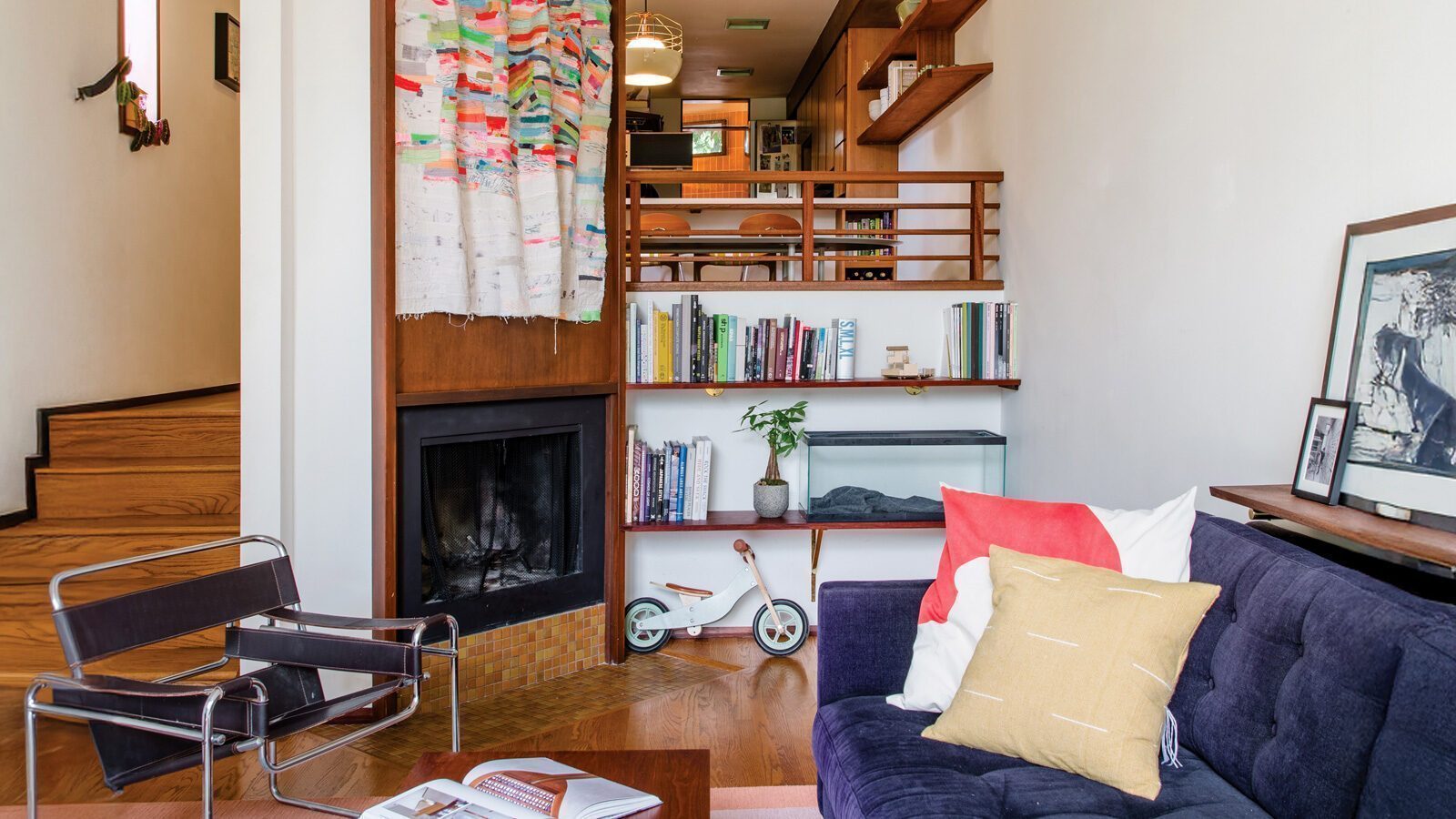 Cozy living room featuring a purple sofa, modern chair, and fireplace. Shelves display books and a small bike. Stairs lead to a dining area partially visible above. Decorative art and pillows add charm to this space, reminiscent of Frank Lloyd-Wright's touch on home renovations by architects.