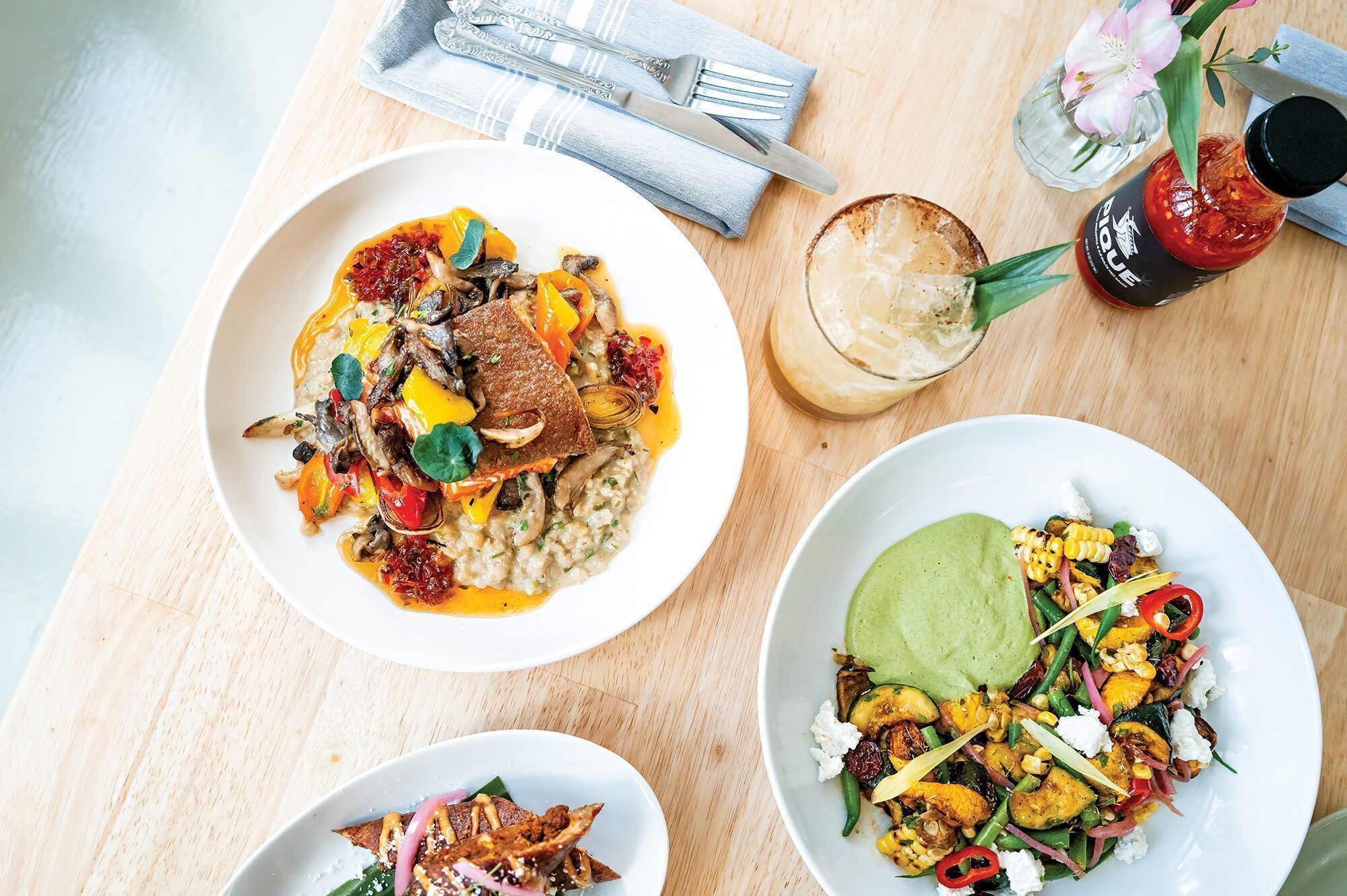 Three plated gourmet dishes by Johnny Reyes, a Lenox beverage with ice and garnish, cutlery, napkin, hot sauce, and a flower are artfully arranged on a light wooden table, viewed from above.