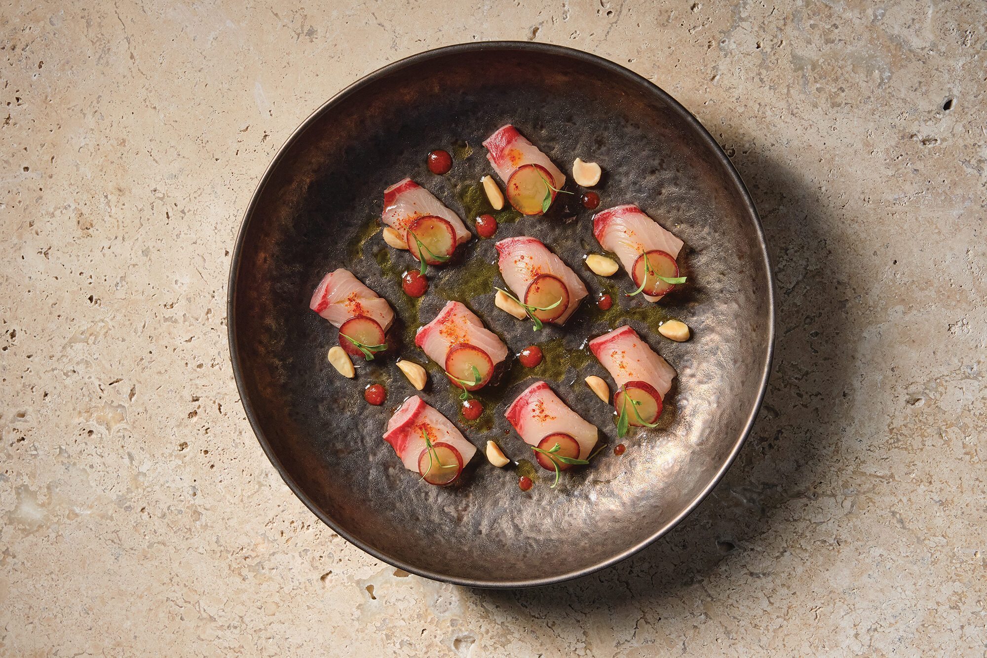 A metal bowl contains nine pieces of sliced fish garnished with almonds, green herbs, red sauce dots, and thin slices of radish—an artful dish you might find while exploring new bars and restaurant openings in Seattle.