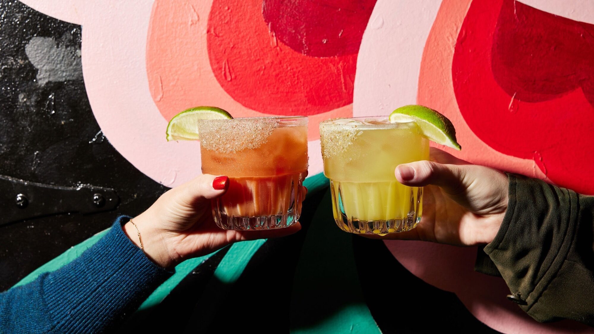 Two hands holding glasses of cocktails with salted rims and lime wedges, set against a colorful abstract mural—perfect for anyone exploring Seattle restaurants or catching up on the latest Seattle food news.