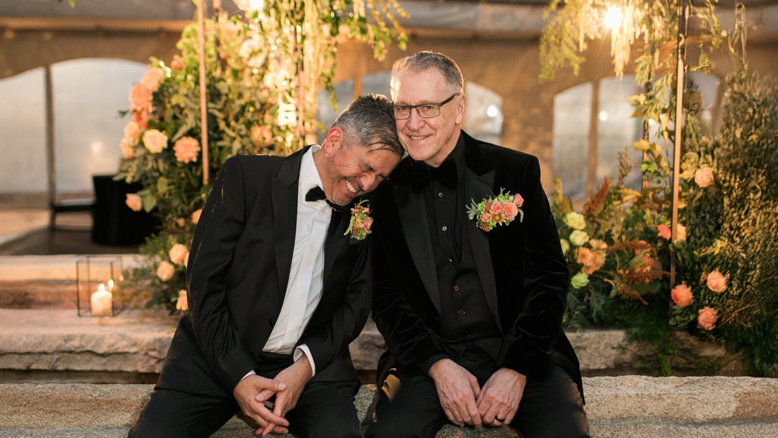 Two men in tuxedos, with floral boutonnieres, sit closely on a stone bench amid floral decorations, appearing content and relaxed at an event.