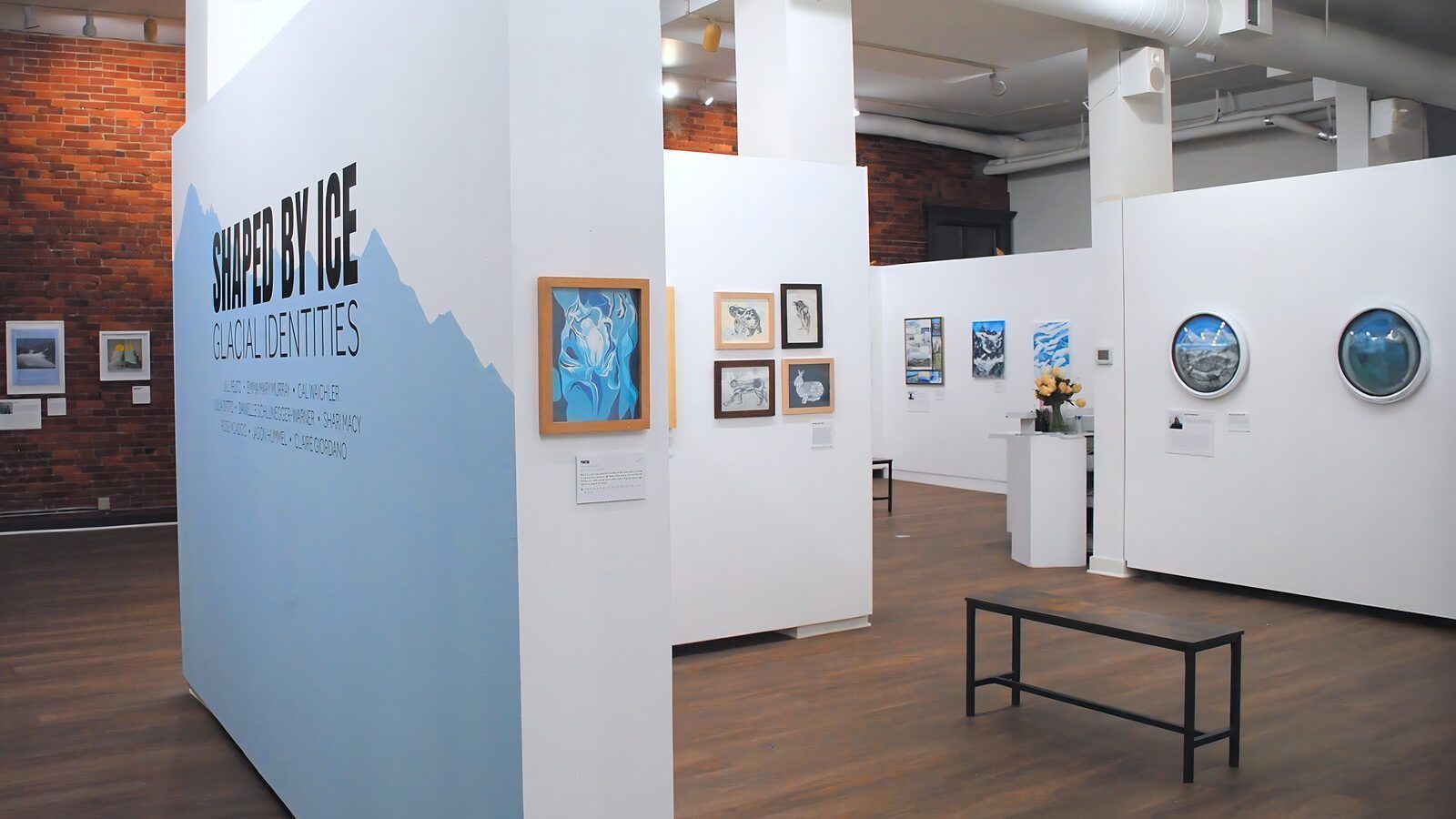 An art gallery exhibit titled "Shaped by Ice: Glacial Expressions" features paintings and framed artwork displayed on white walls with wooden flooring and a black bench.