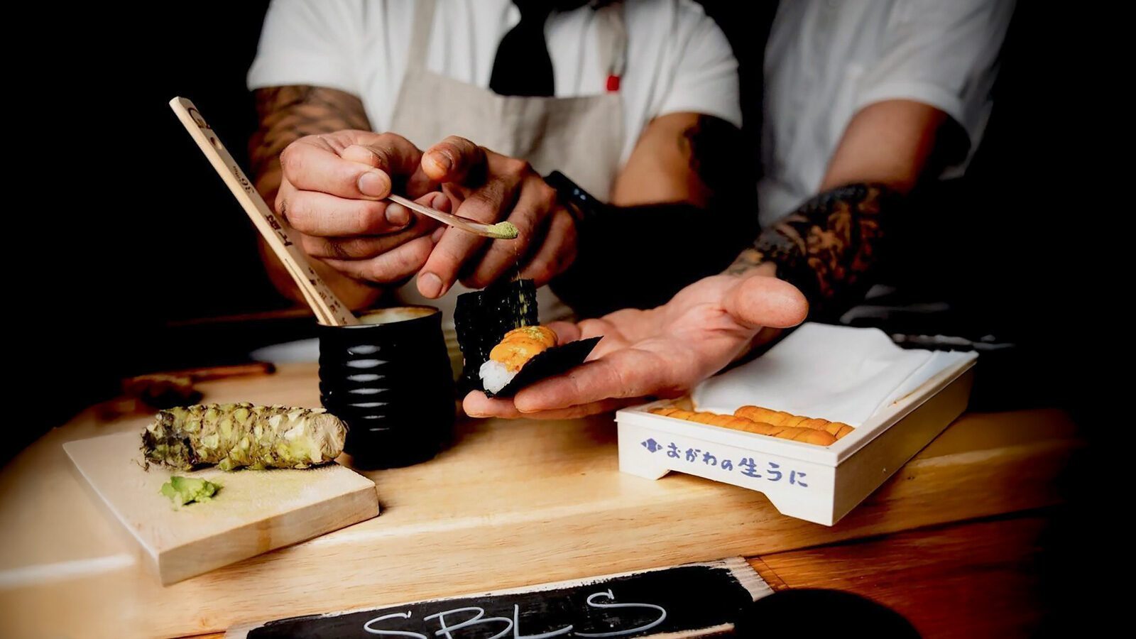 Chef placing wasabi on sushi while another person holds it, capturing a moment worthy of the latest food news. A whole wasabi root and box of sushi sit enticingly on the counter, adding to the vibrant culinary scene reminiscent of a Restaurant Roundup.