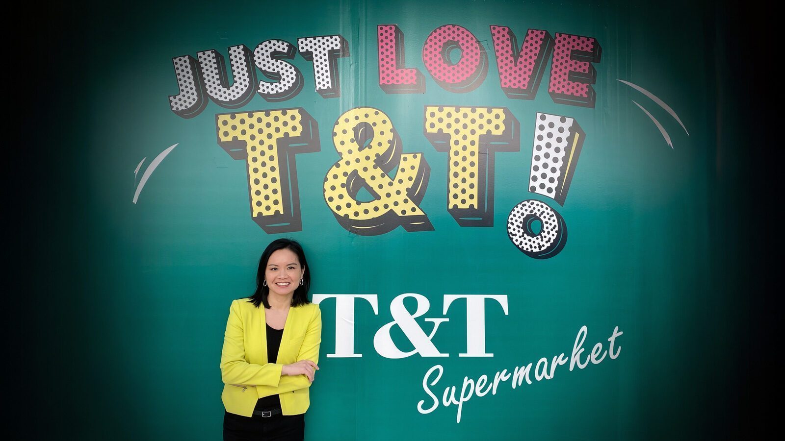 A woman in a yellow blazer stands in front of a green wall with large text reading "JUST LOVE T&T!" and "T&T Supermarket," making it a bright spot featured in Seattle food news and popular among Seattle restaurants.
