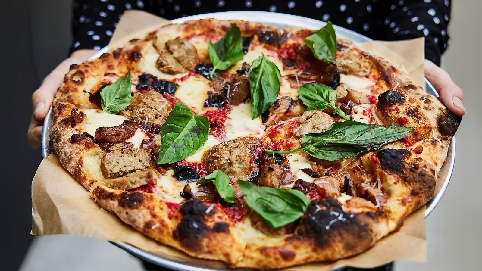 A person holds a round pizza on parchment paper, topped with basil leaves, sausage, cheese, and tomato sauce on a slightly charred crust—just one of the mouthwatering creations Hidden in Plain Sight at The Local 104.