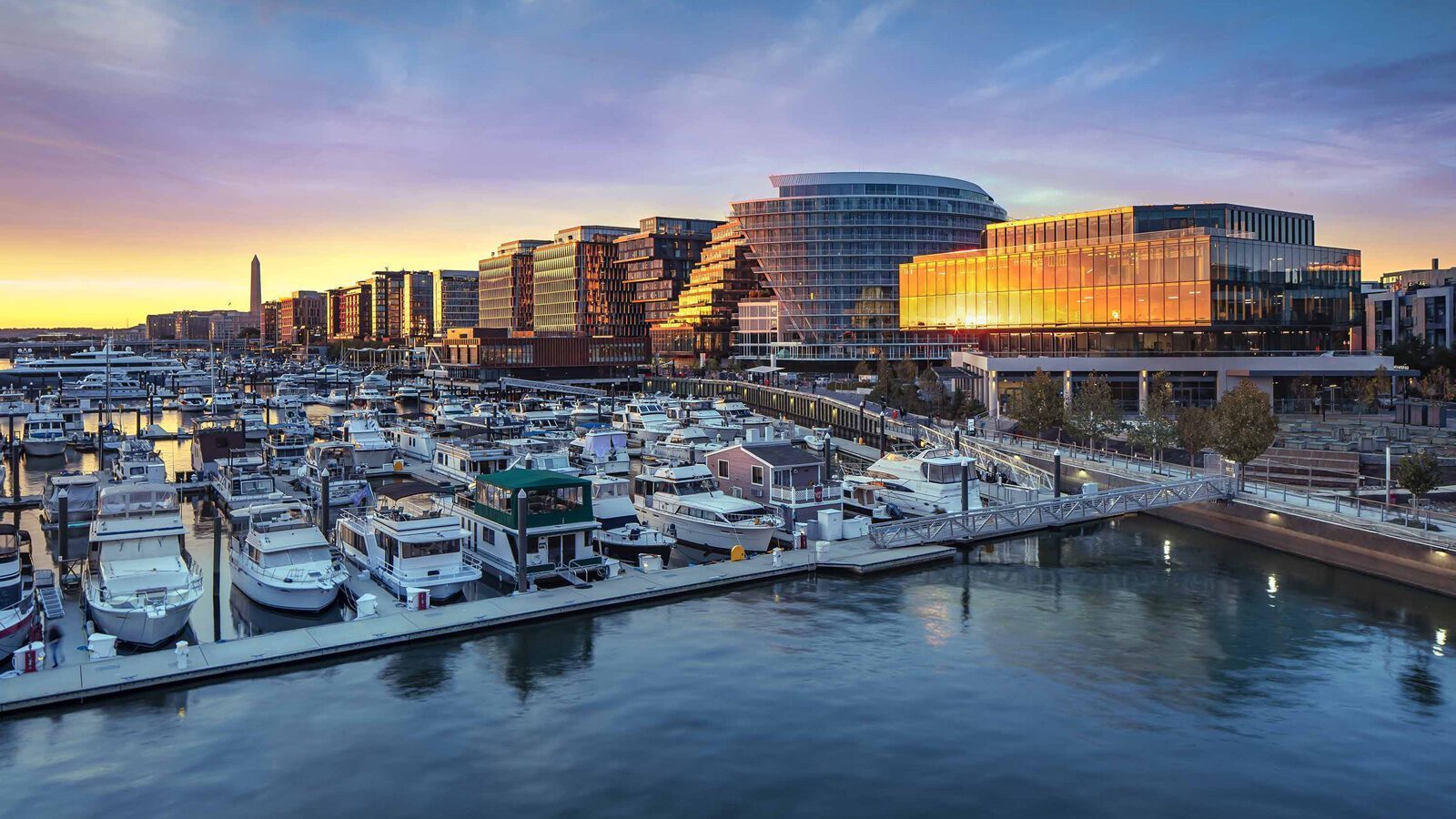 Modern waterfront buildings and a marina with docked boats at sunset reflect light on the water—capturing the essence of A Stroll Through Old & New D.C. against an urban skyline in the background.