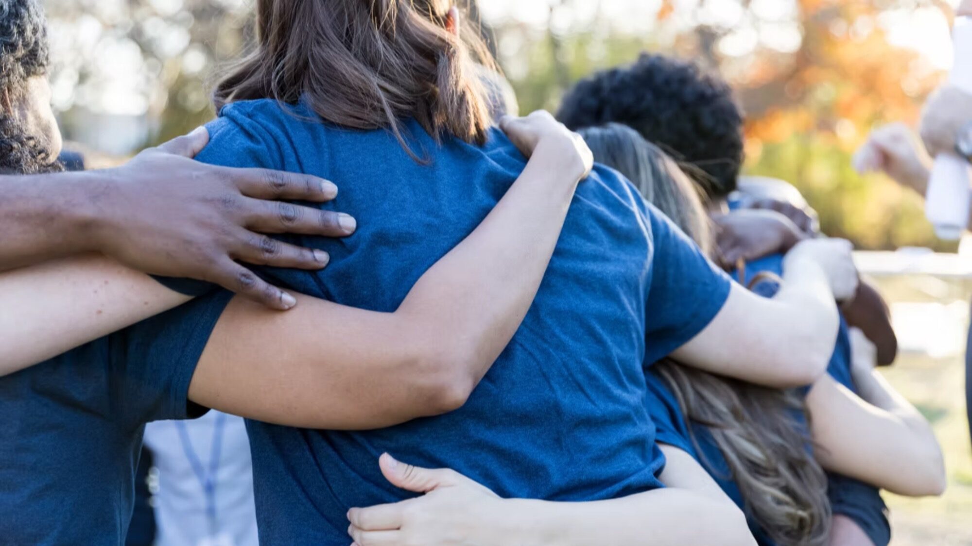 A group of people wearing blue shirts stand outdoors with their arms around each other, embodying the spirit of camaraderie fostered by Eastside Youth Services.