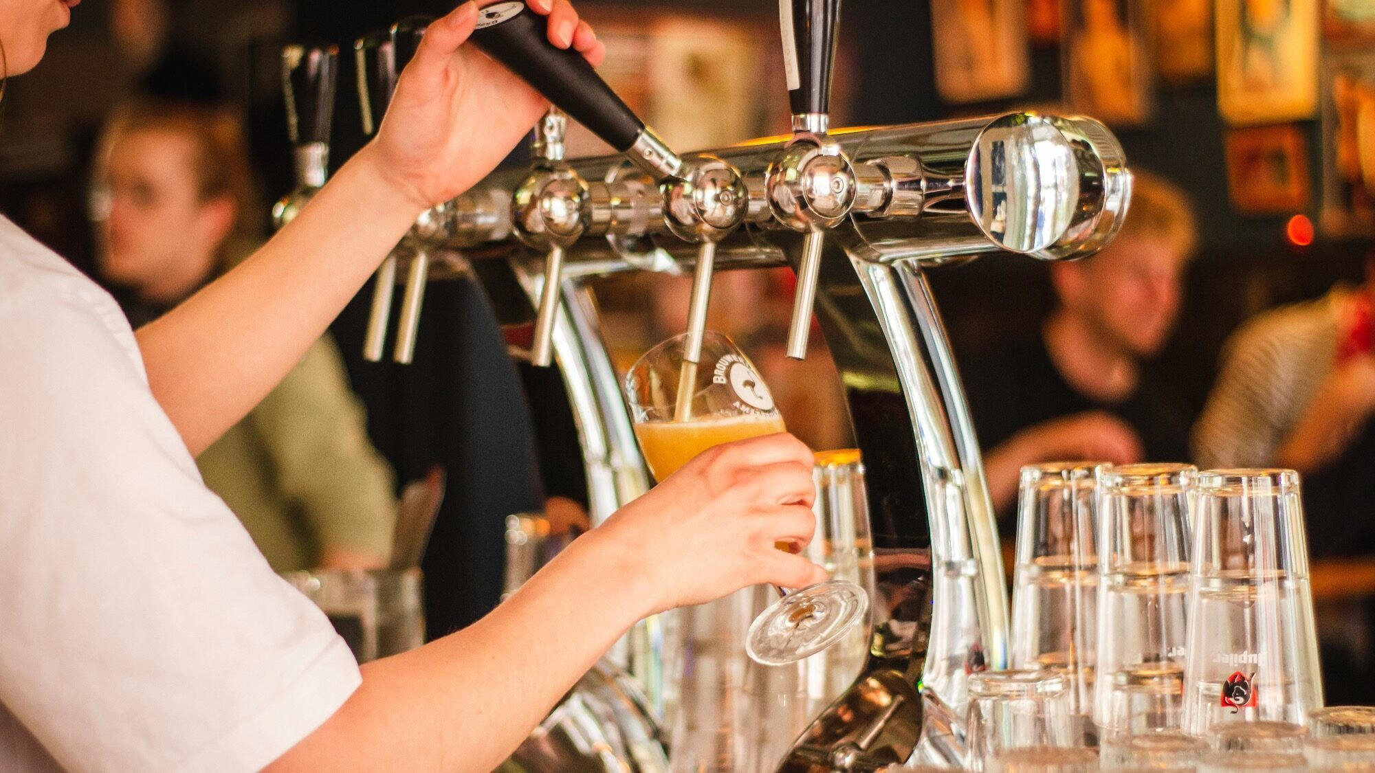 A person pours beer from a tap into a glass at a bar, with stacked glasses and blurred people in the background.