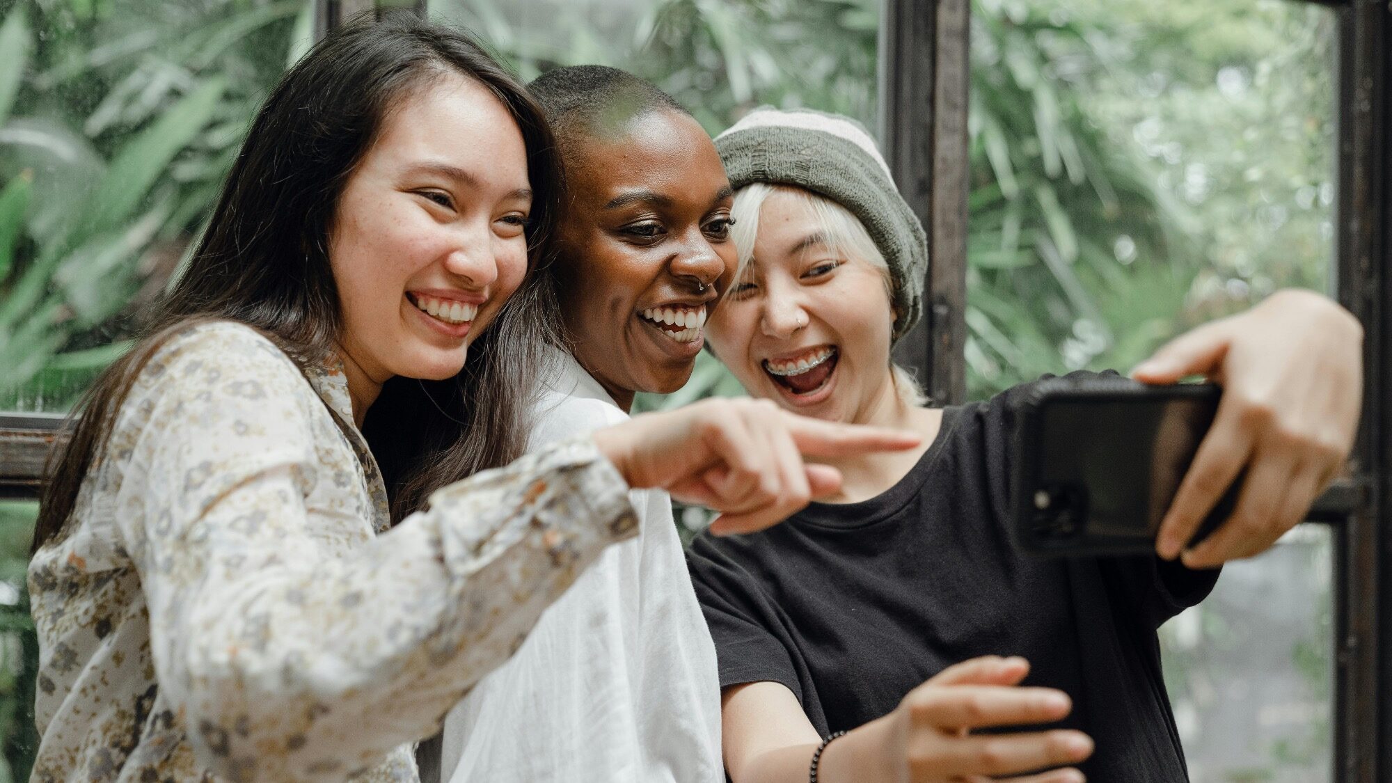 Three people stand close together, smiling and pointing at a smartphone while taking a selfie indoors with greenery visible through the window behind them.