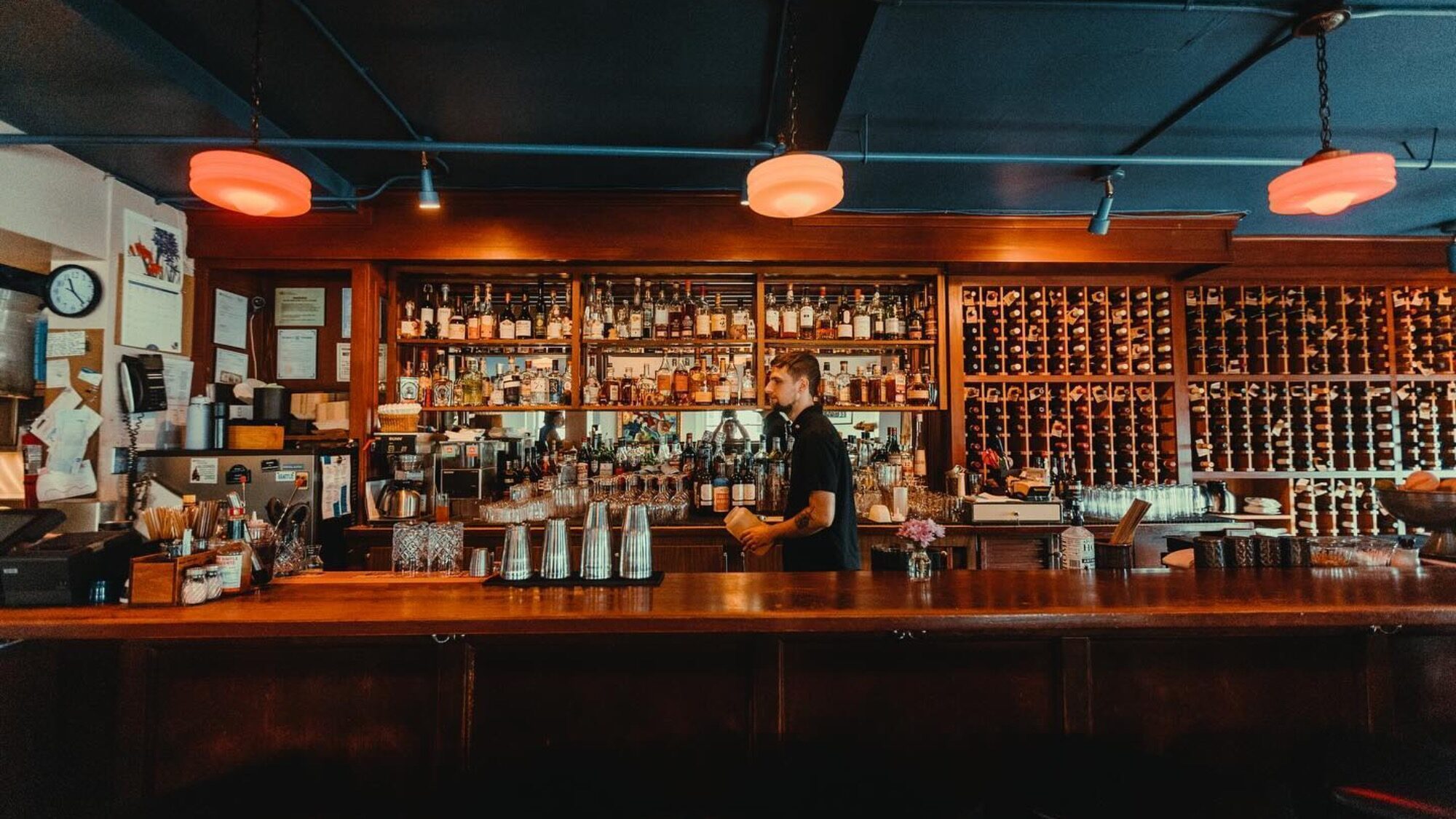 A bartender stands behind a wooden bar counter with shelves of liquor bottles and wine in the background, under warm ceiling lights.