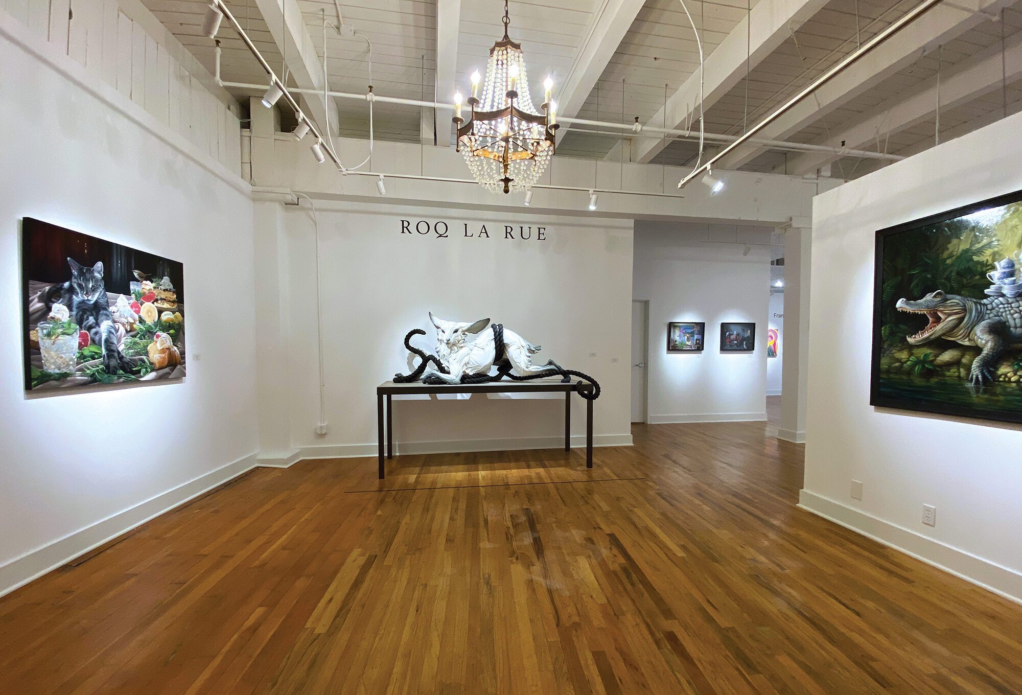 Art gallery interior with wooden floors, featuring paintings of animals on the walls and a sculpted white cat with tentacles, displayed centrally under a chandelier—evoking the unique aesthetic of gallerist Kirsten Anderson's Roq La Rue art space.