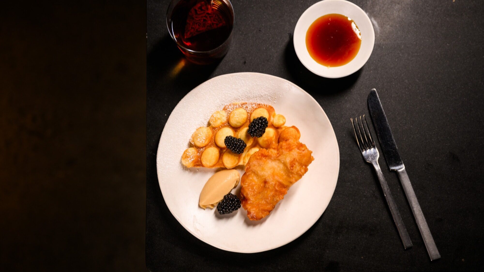 A white plate with waffles, a scoop of ice cream, berries, and a piece of fried chicken. A cup of syrup and a drink are nearby, with a knife and fork placed on the right.