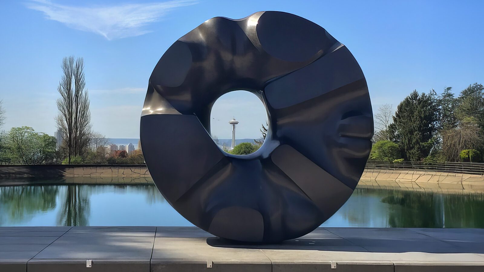 Large black abstract circular sculpture with a central hole, displayed outdoors near a reflective pool, stands among trees and a tower—one of the striking Seattle cultural landmarks.