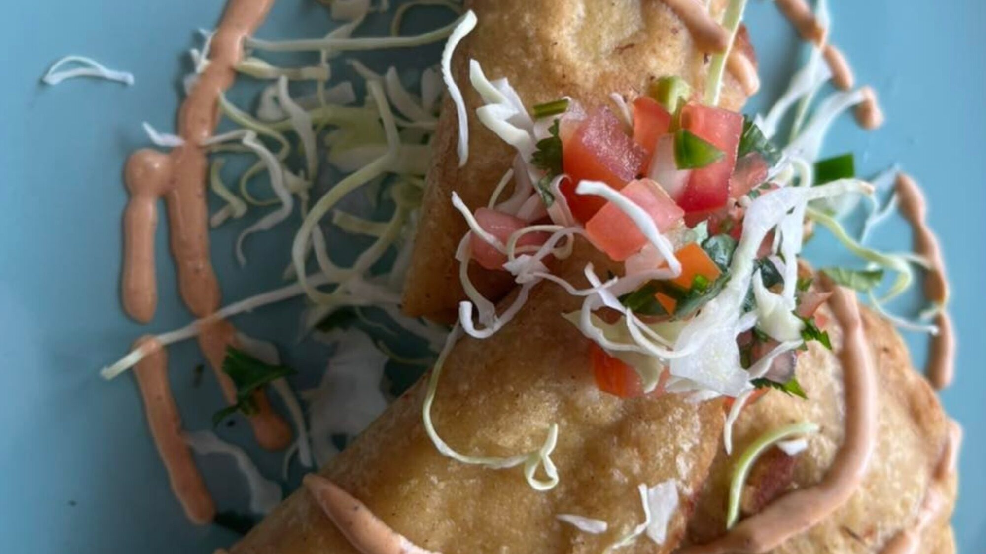Two fried tacos topped with shredded cabbage, diced tomatoes, onions, and cilantro, drizzled with a creamy sauce, served on a blue plate.