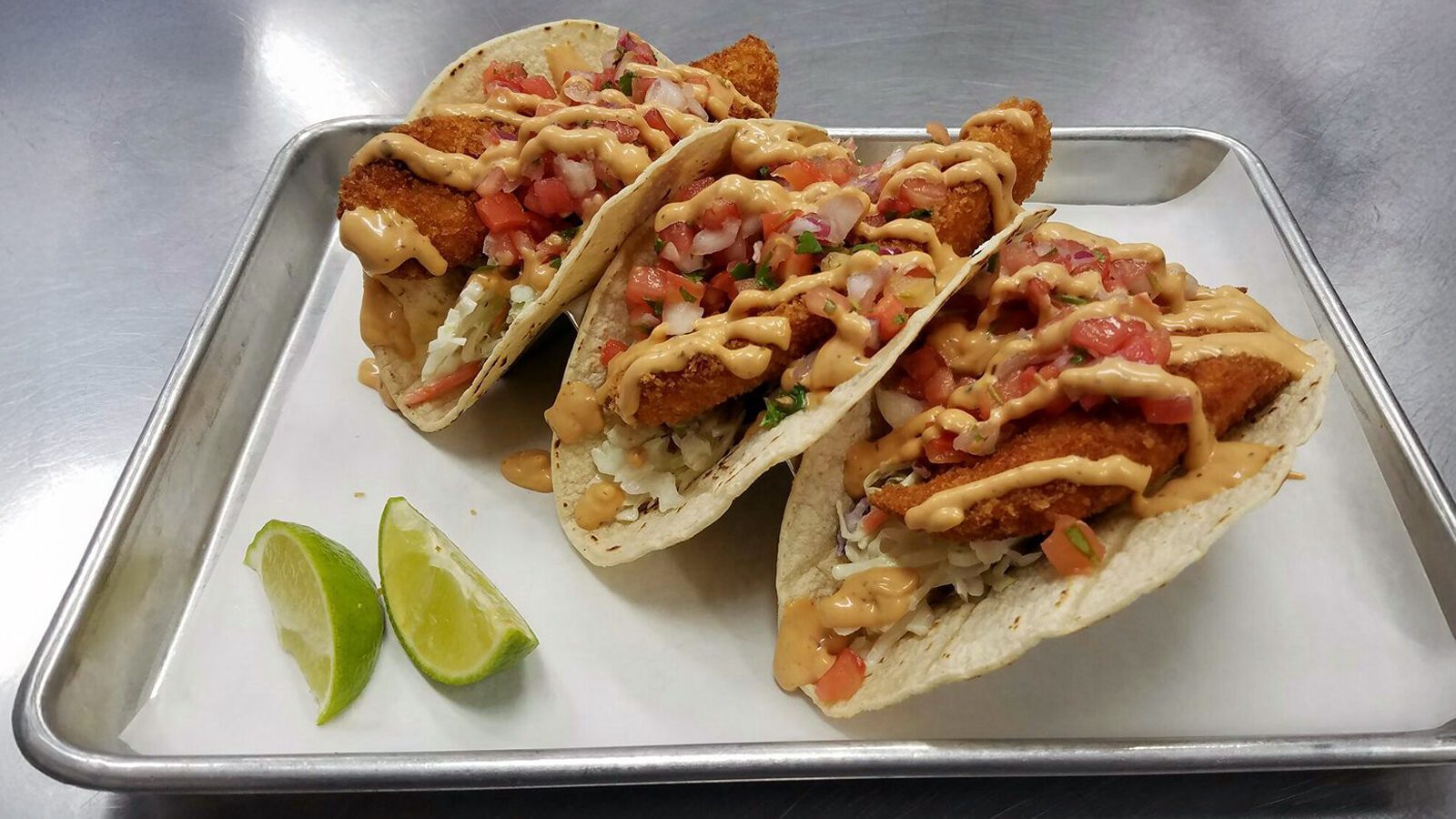 Three breaded fish tacos from Counter Culture: Burien Fish House, topped with diced tomatoes, shredded cabbage, and creamy sauce on tortillas, served on a metal tray with two lime wedges.