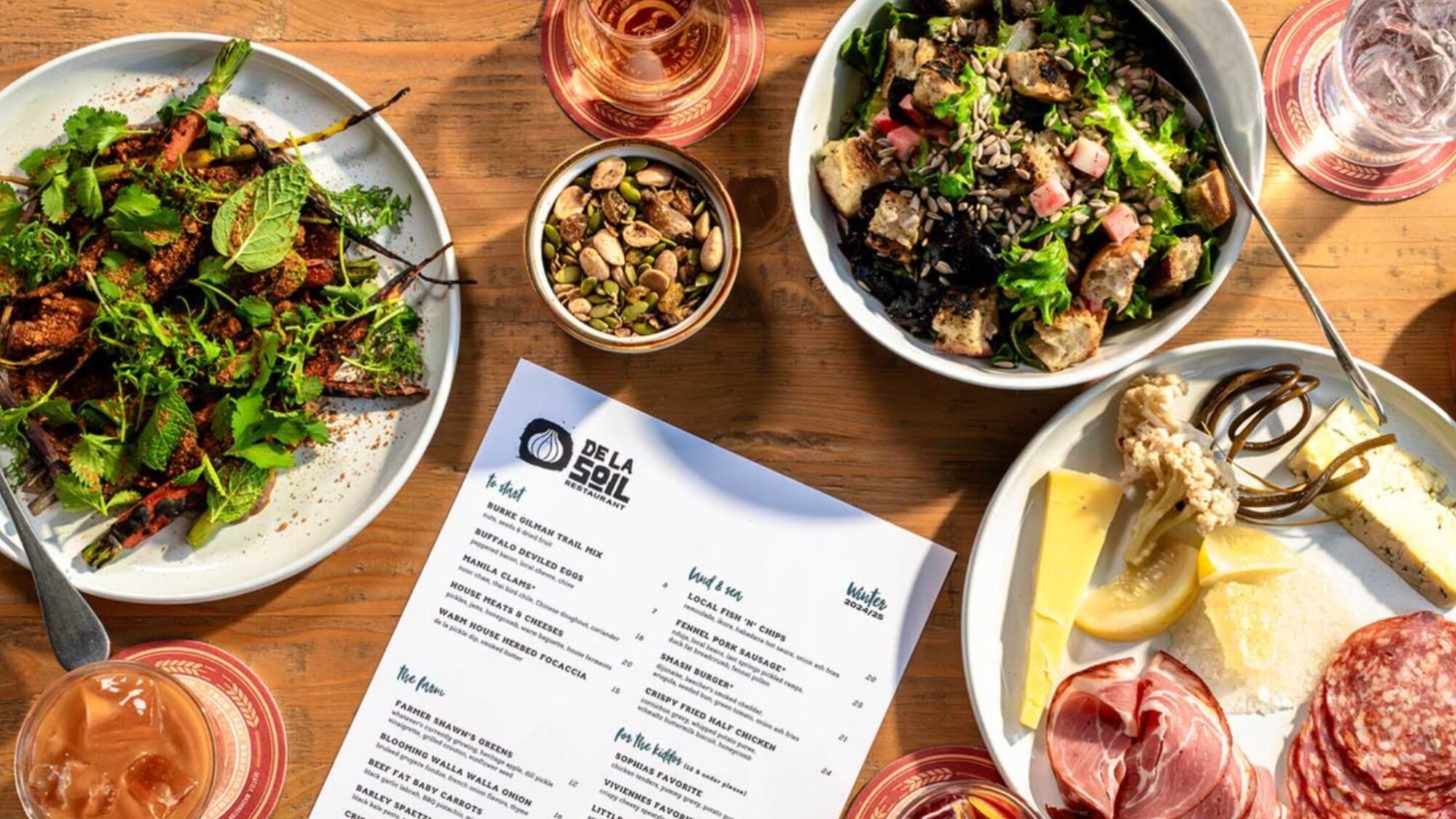 Overhead view of a wooden table with assorted dishes, including salad, roasted vegetables, charcuterie, nuts, cheese, and a restaurant menu in the center.