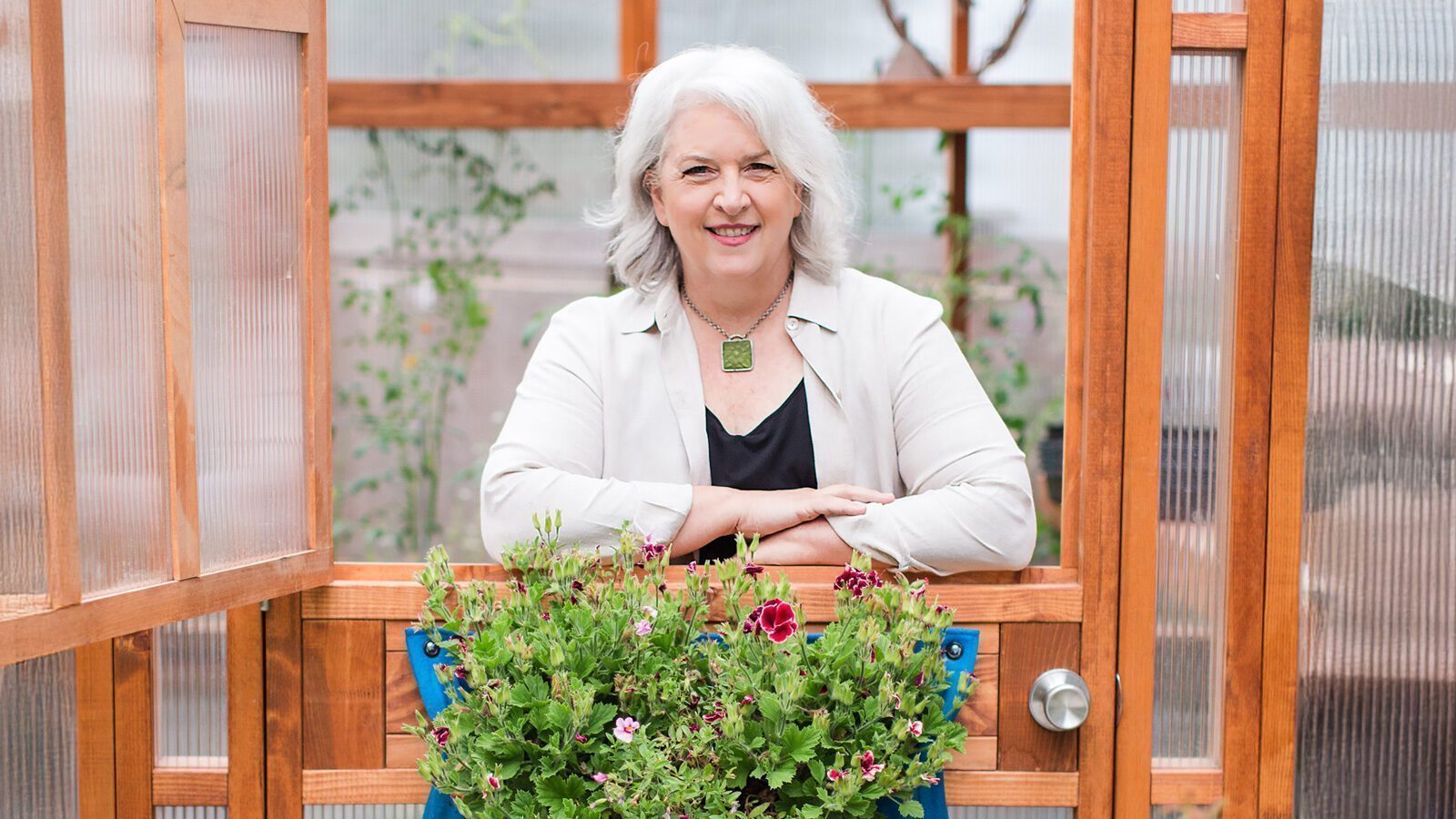 An older woman with gray hair stands smiling behind a wooden door, with blooming local flowers in front of her inside a greenhouse—a scene reminiscent of Debra Prinzing's focus on seasonal and sustainably harvested flowers.