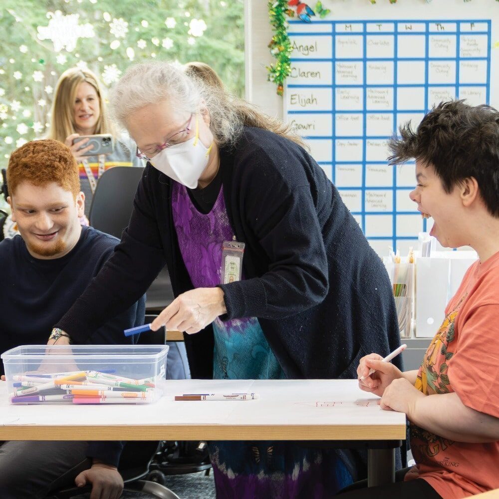 Adults from the Evergreen Transition Program, including students with disabilities, sit and stand around a table working on an art project with markers in a classroom setting.person