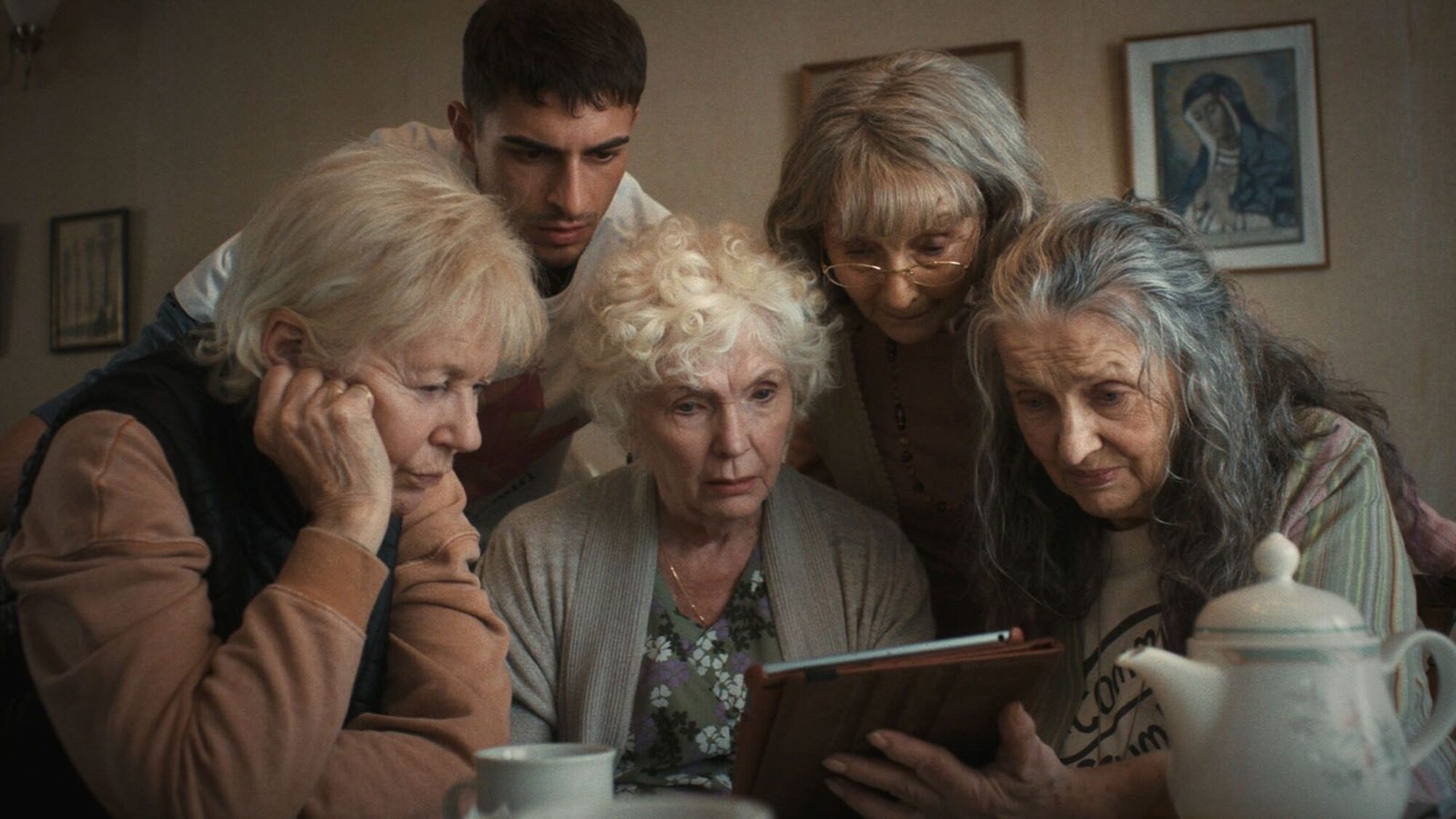 Five elderly women and a younger man closely look at a tablet together around a table in a cozy, softly lit room with framed art on the walls.