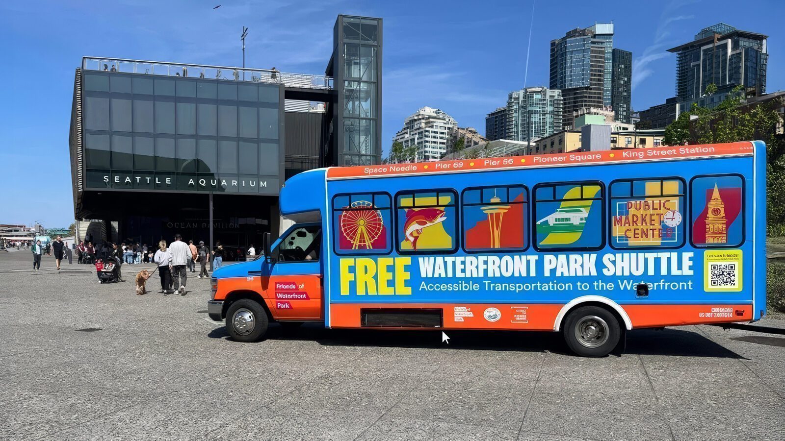 A brightly colored Free Waterfront Shuttle labeled "Free Waterfront Park Shuttle" is parked outside the Seattle Aquarium, with city buildings visible in the background.