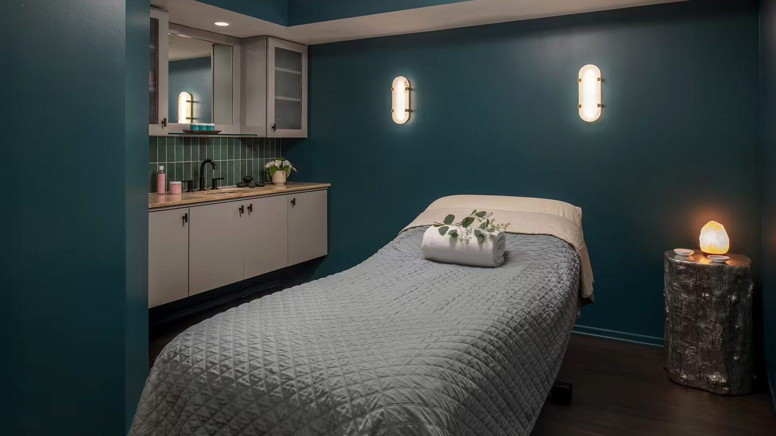 Treat Mom to some Emerald City Elegance this Mother’s Day in a tranquil massage room featuring a neatly made bed, rolled towel with leaves, soothing wall lights, a sink with cabinets, and a glowing salt lamp on a small table.