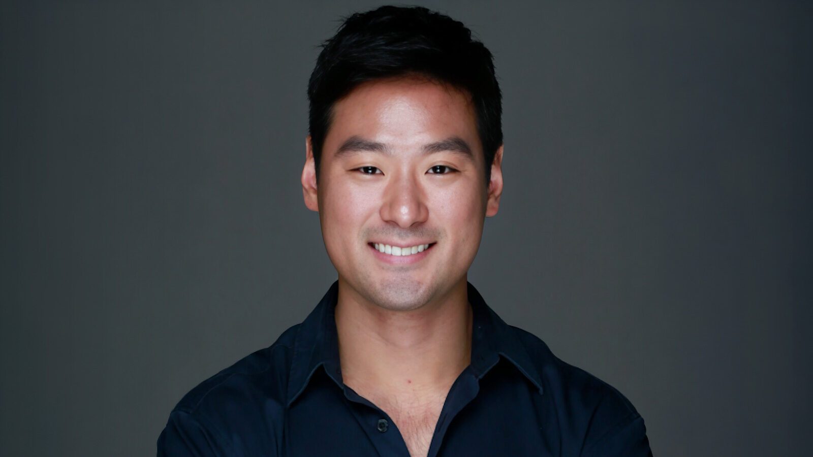 A man with short black hair and a dark shirt smiles at the camera against a plain dark background—Music Producer Kyu Lee talks Seattle, UW, and his Hollywood career.