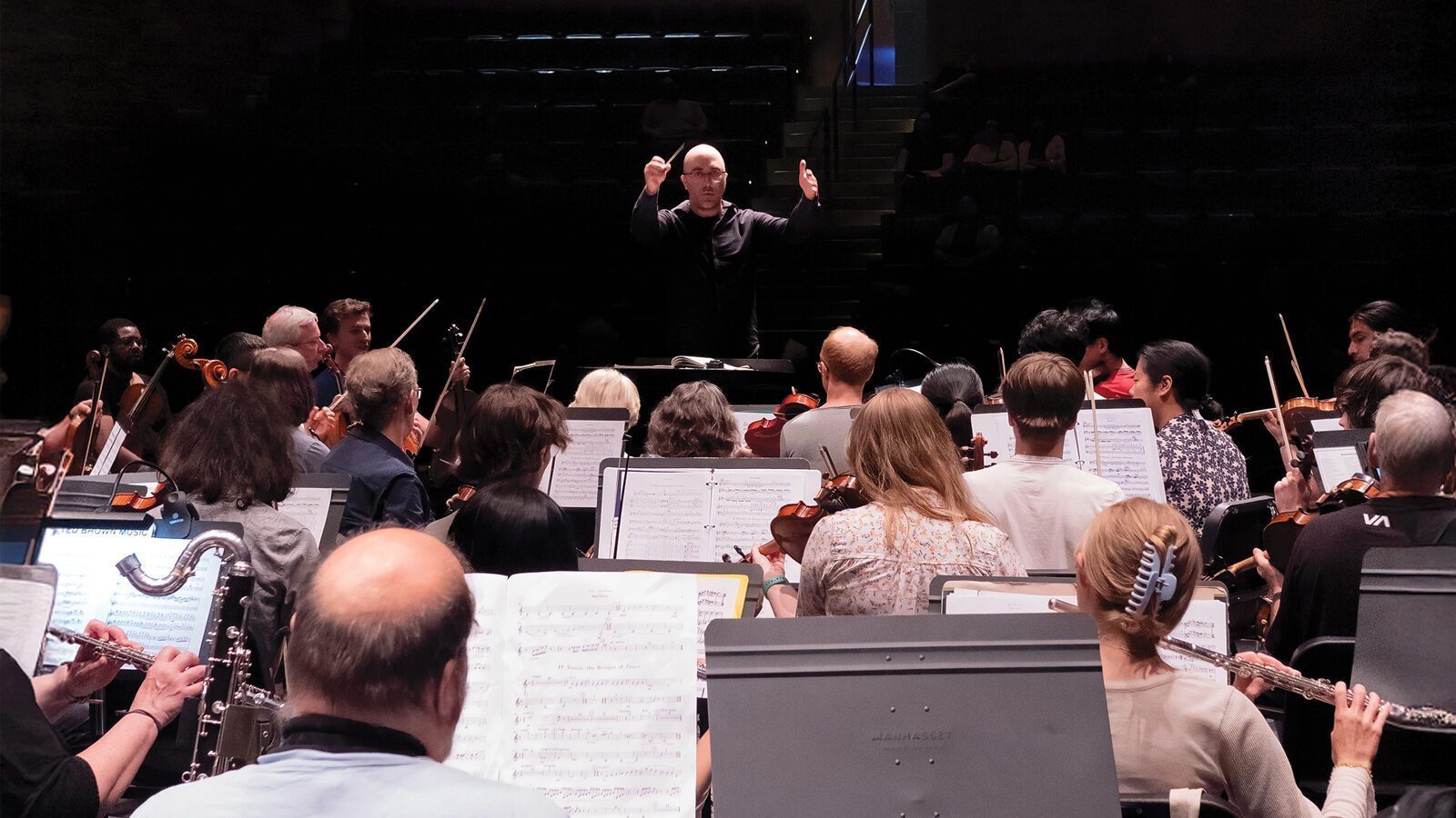 A conductor leads an orchestra during a rehearsal at the Shorewood Performing Arts Center, with musicians reading sheet music beneath dim lights as they prepare for the Mahler Fest 30th anniversary.