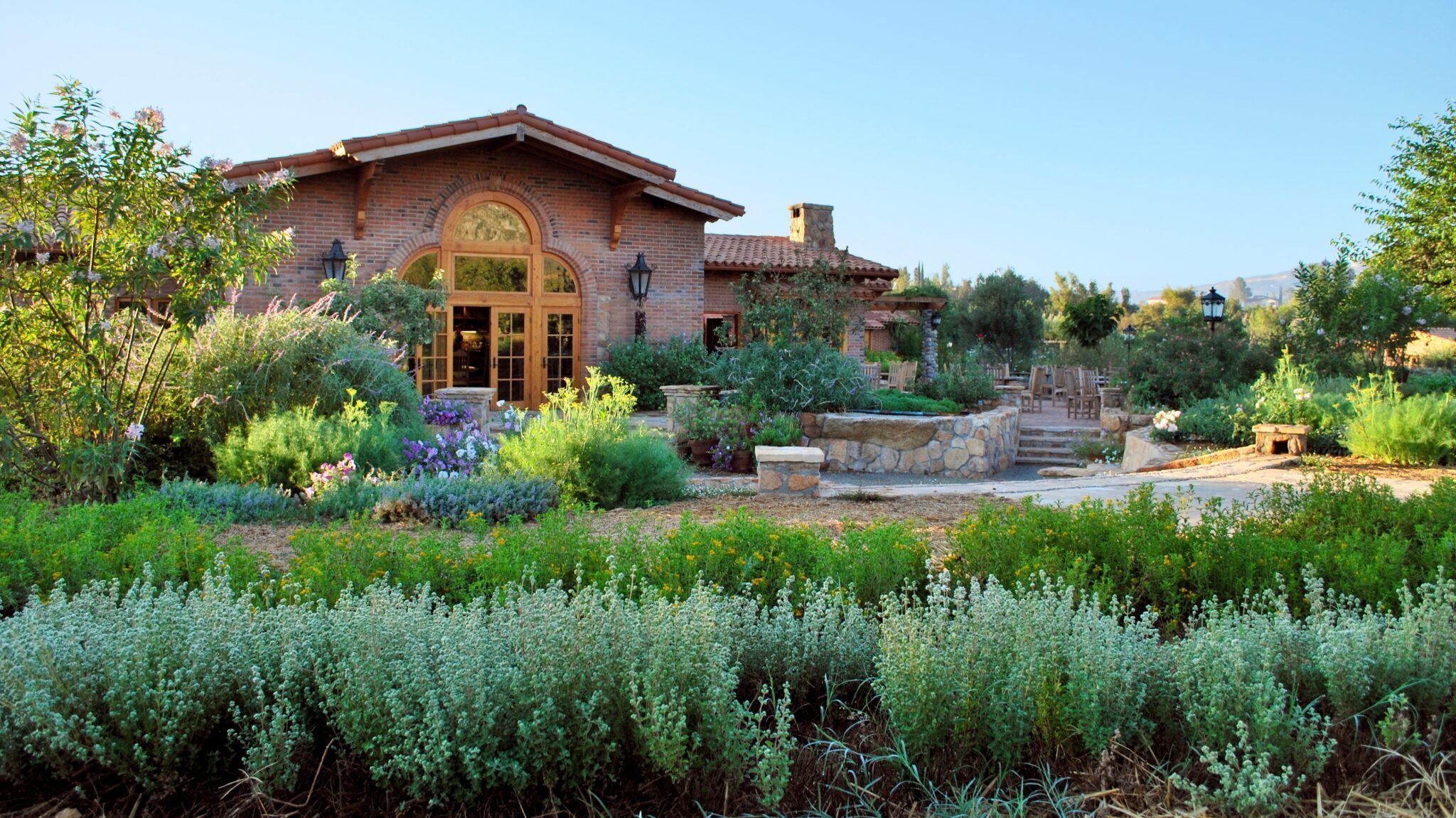 A rustic house with an arched window is surrounded by lush gardens, shrubs, and stone pathways under a clear blue sky, creating a sanctuary reminiscent of Rancho La Puerta.