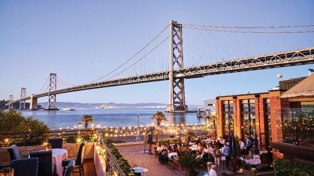 Outdoor restaurant diners in San Francisco sit near the waterfront at sunset with string lights, many wearing trendy sneakers, as a large suspension bridge provides a stunning backdrop.