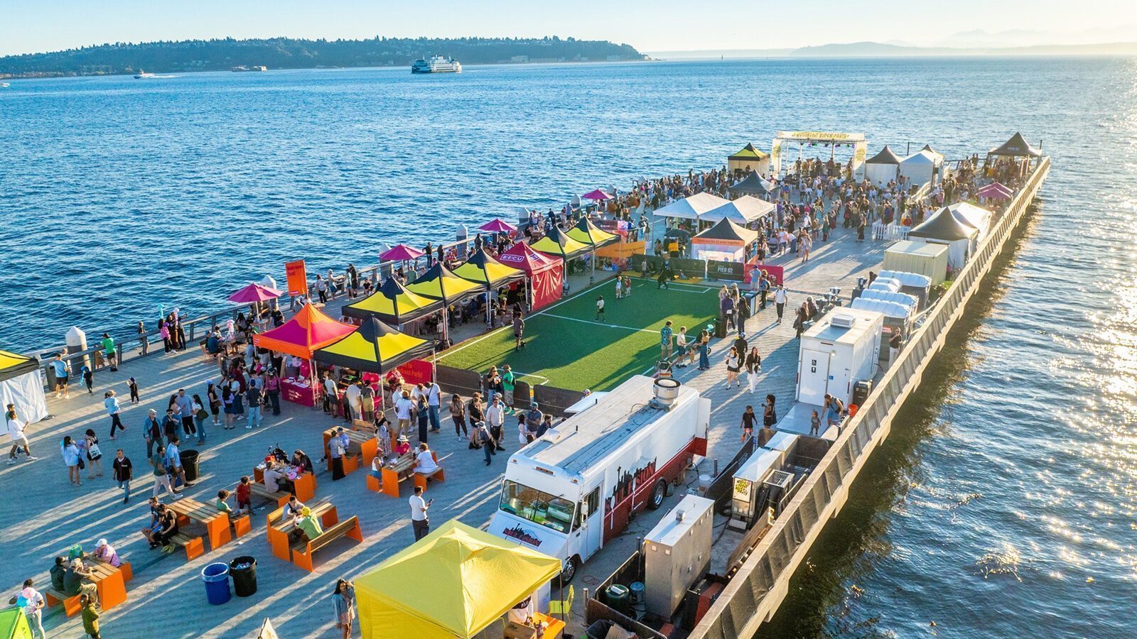 Aerial view of a busy pier with colorful tents, food trucks, and crowds enjoying Seattle sports by the water on a sunny day.