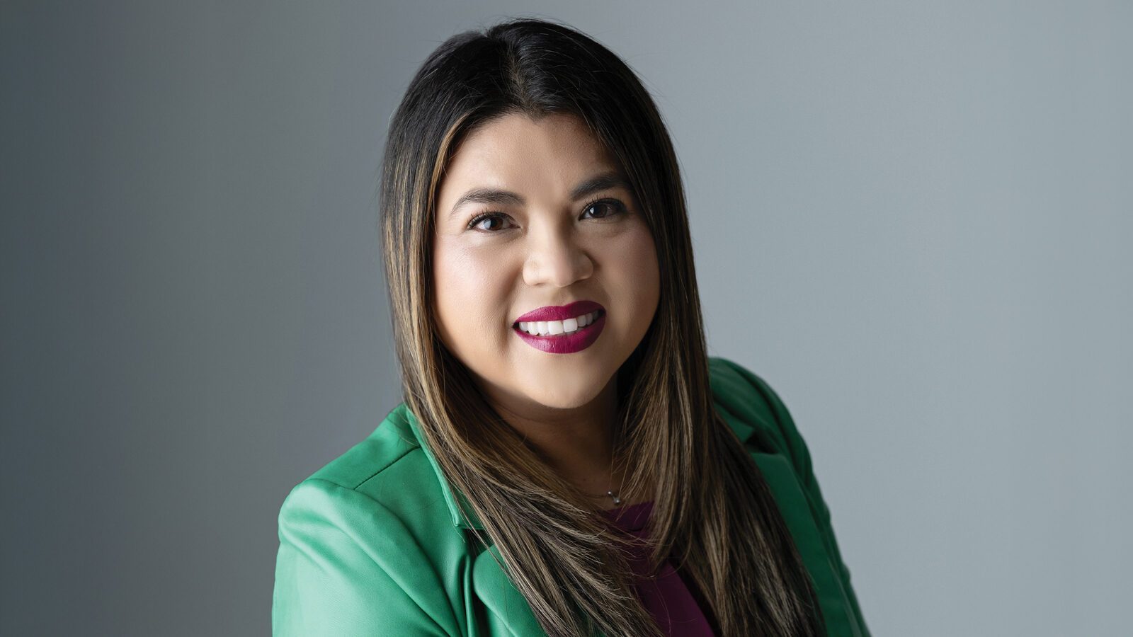 Jaqueline Garcia Castillo, a Latina woman with long, straight brown hair, wears a green blazer and maroon top, smiling confidently in front of a plain gray background.