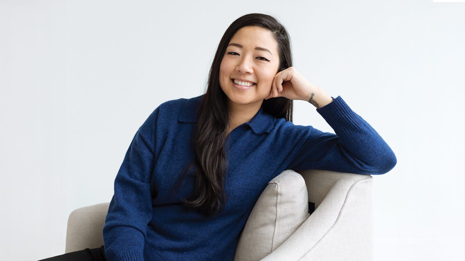 Joan Sung, a Korean American woman, with long dark hair and a blue sweater, sits on a light-colored chair, smiling with her head resting on her hand against a pale background.