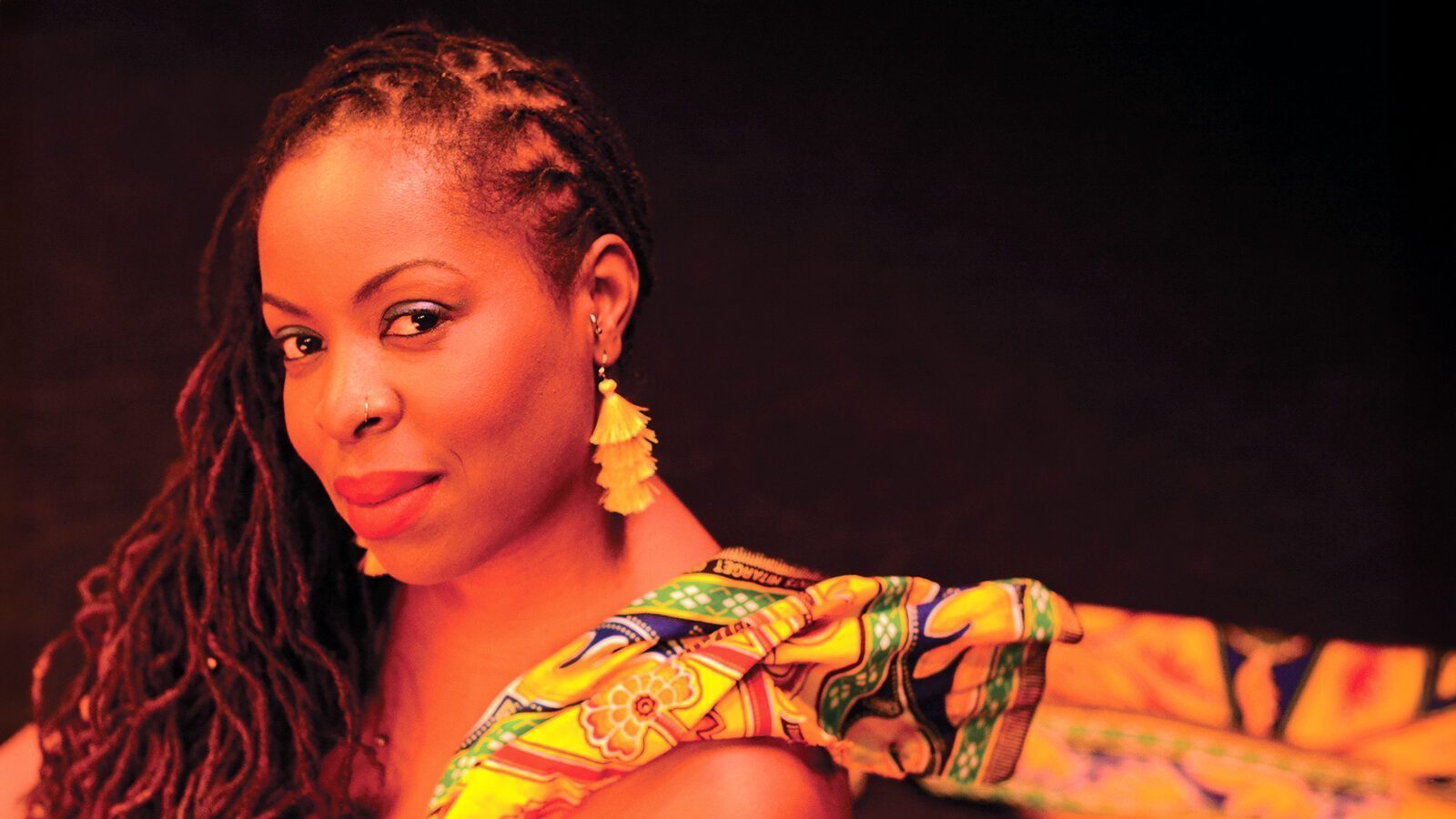 A woman with long, braided hair and yellow tassel earrings looks at the camera, exuding artsy flair in a colorful patterned garment against a dark background—reminiscent of jazz musician Johnaye Kendrick’s vibrant style.