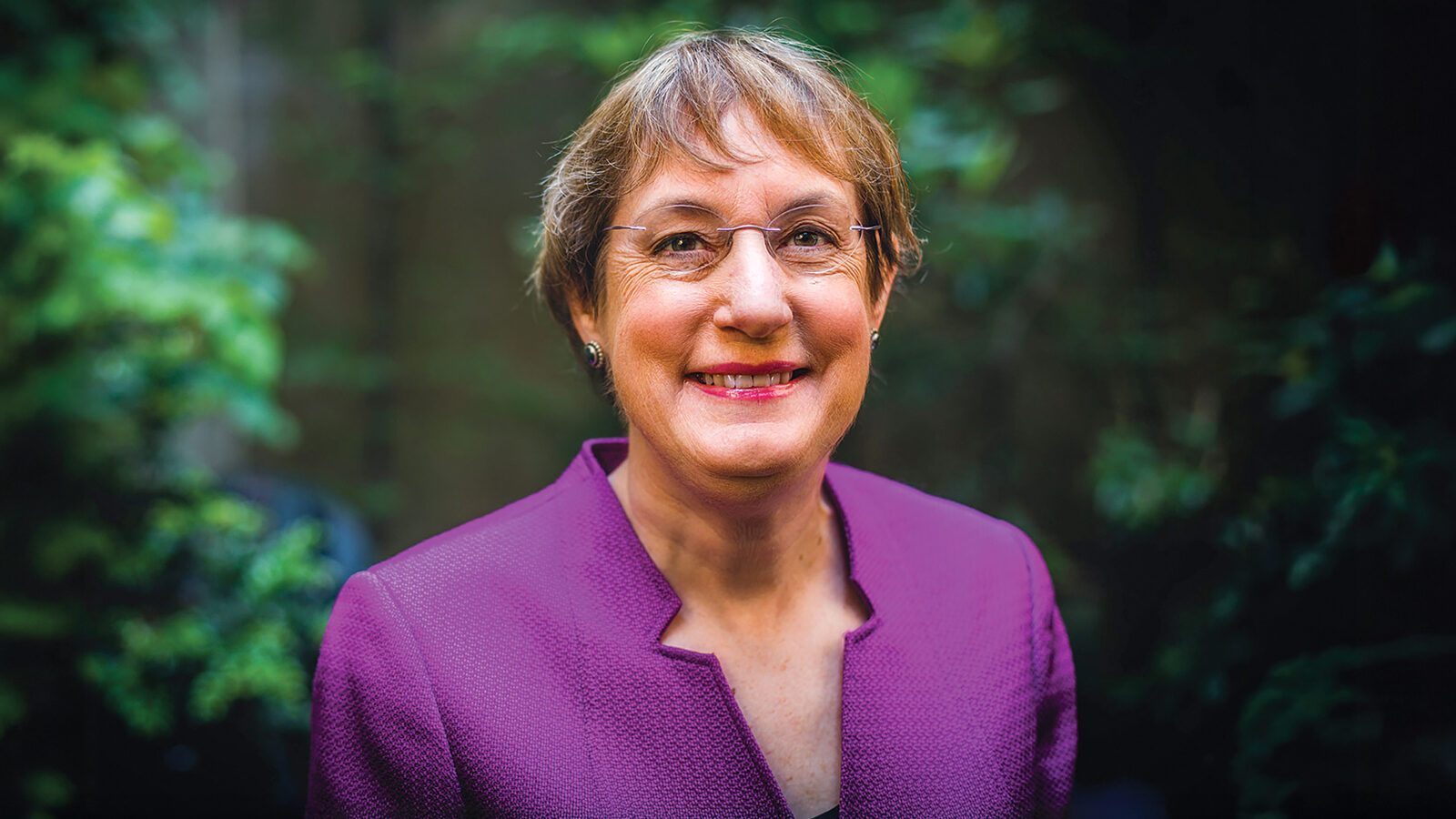 A middle-aged woman with short light brown hair, glasses, and a purple blazer smiles outdoors in front of leafy green foliage, celebrating women in science.