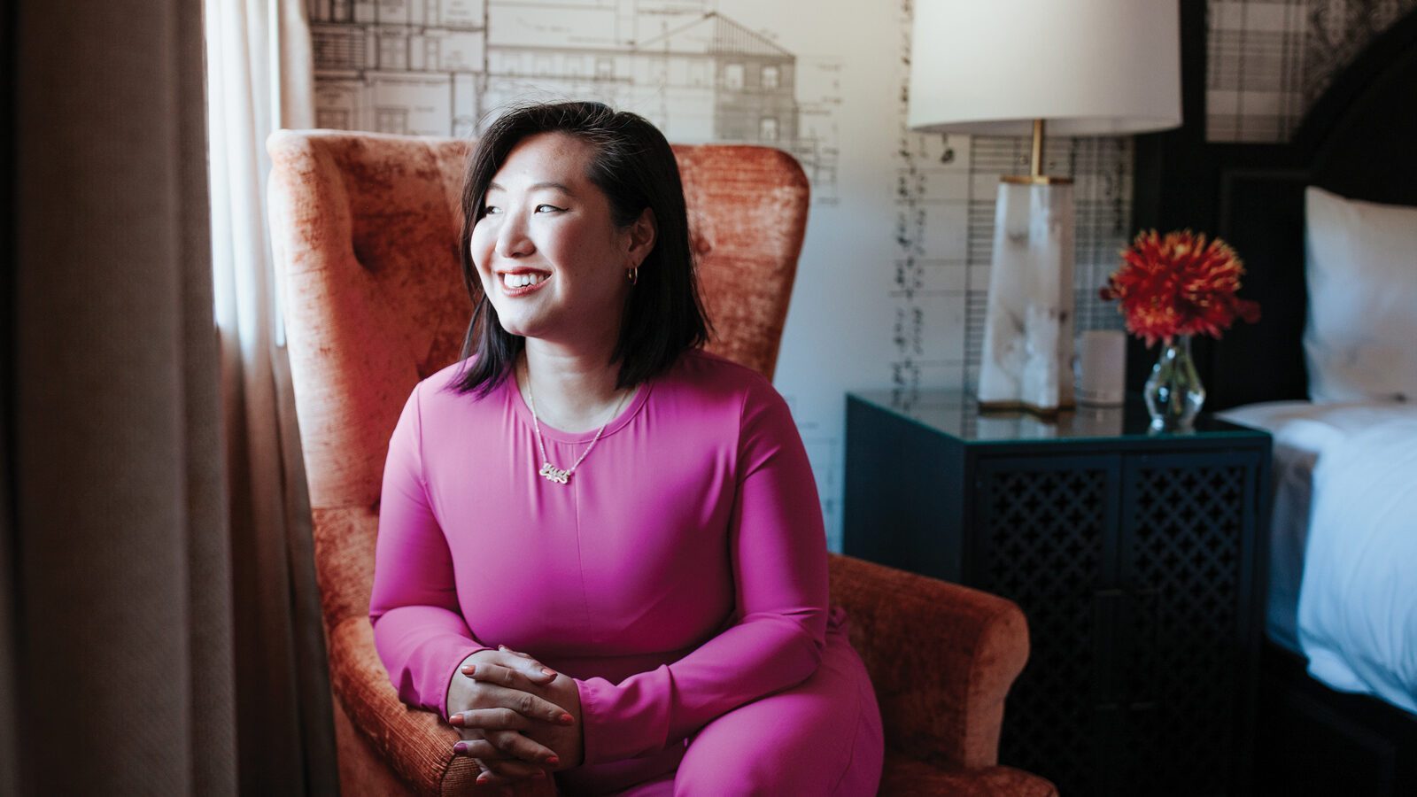 Trailblazing Women: Stephiney Foley sits in a bright pink outfit on an orange chair, smiling as she looks toward the window. A lamp and a vase with red flowers adorn the table beside her.