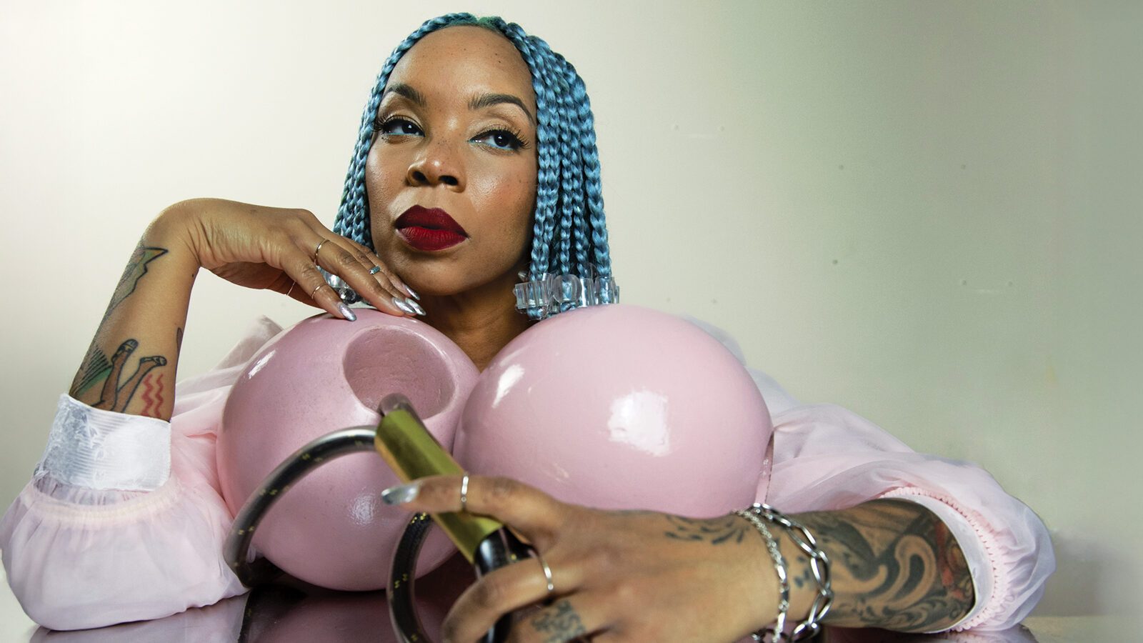 Trailblazing Women: A person with blue braids and red lipstick poses with their hand resting on two large pink spherical objects and a brass handle, displaying tattoos and silver jewelry in a style reminiscent of Tariqa Waters.