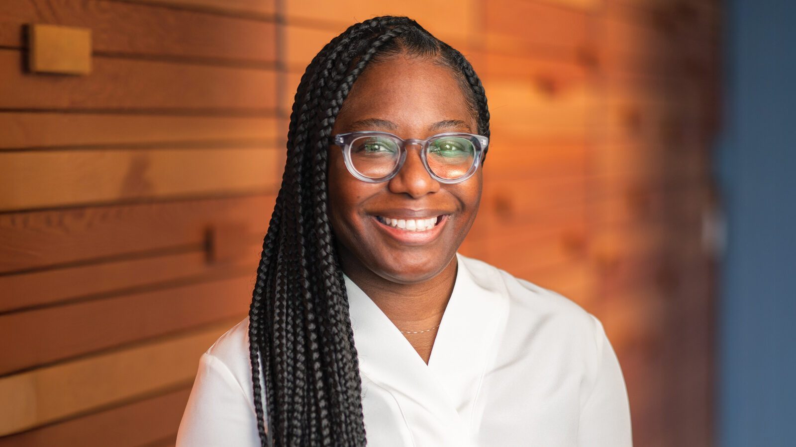A woman of color with long braided hair, glasses, and a white top smiles at the camera while standing in front of a wooden wall.