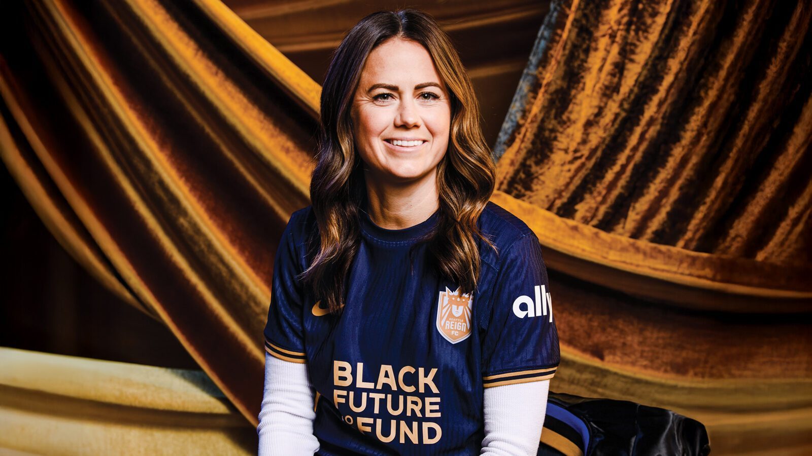 Lauren Barnes, a woman with long brown hair, smiles while wearing a navy blue Seattle Reign FC soccer jersey with "Black Future Co-op Fund" text, seated in front of gold draped fabric.